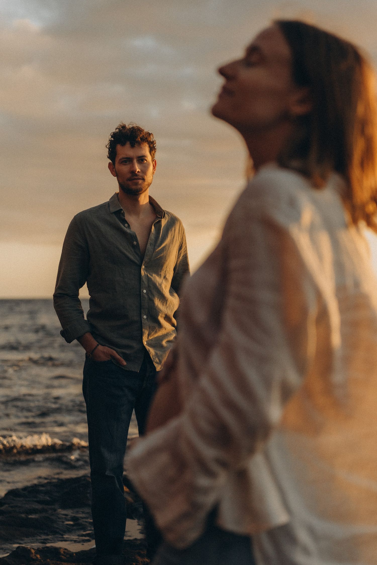 Couple at sunset on rocky beach, man in grey shirt gazes at woman in white blouse with golden light.