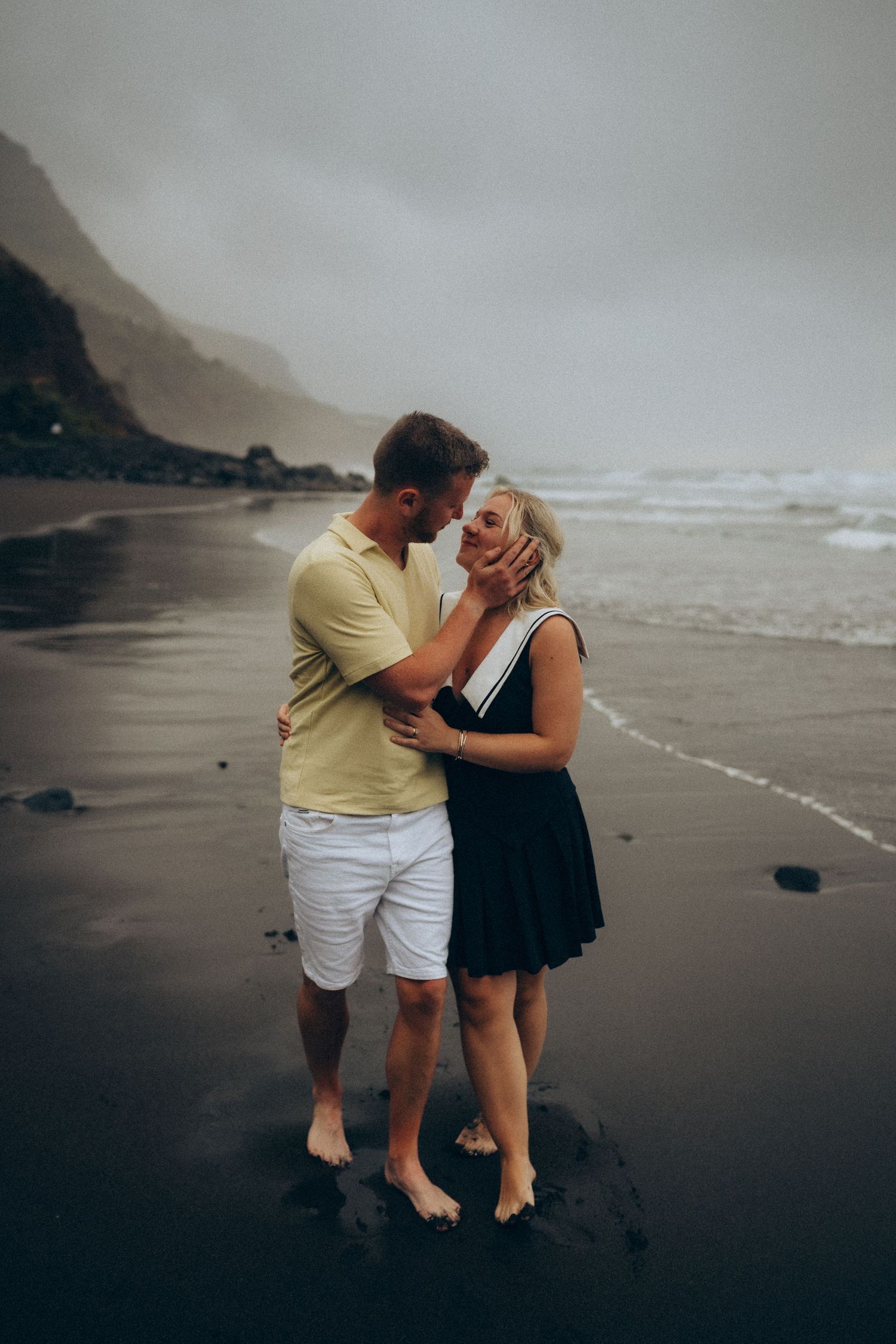 Couple sharing a romantic moment on a dark volcanic beach with misty mountains and ocean waves in the background.