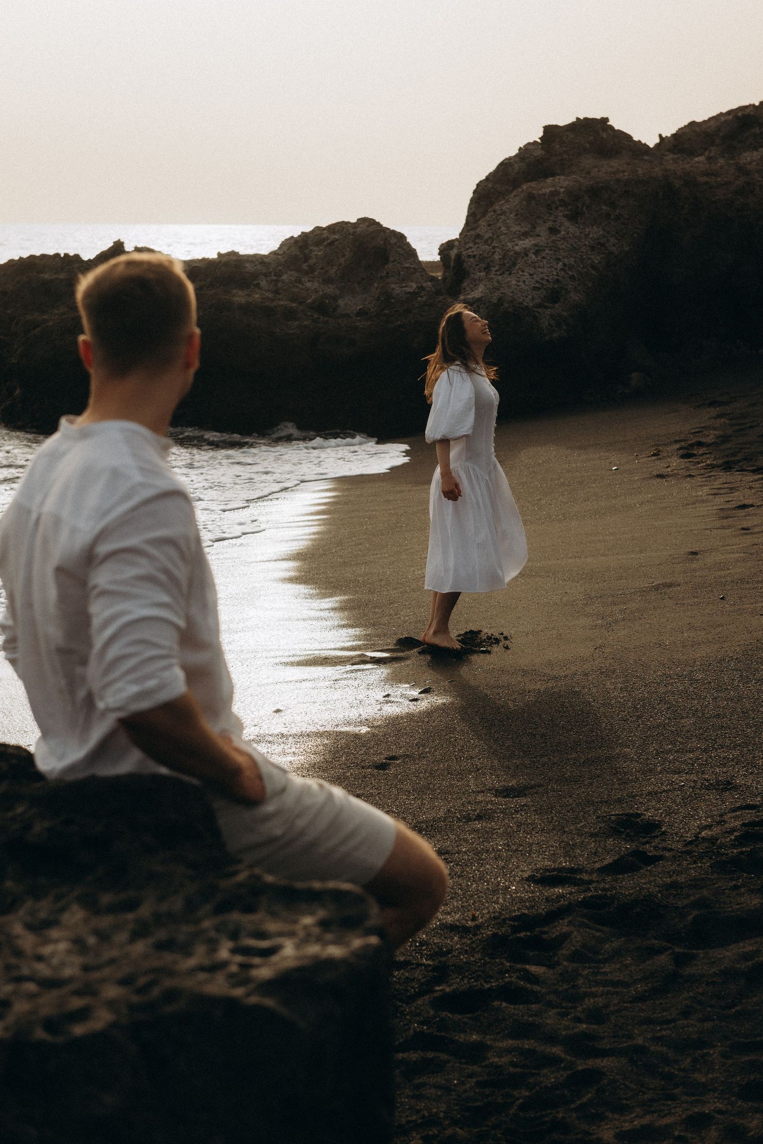 A couple in white clothing on a rocky beach at dusk, woman standing near water as man watches from rocks.
