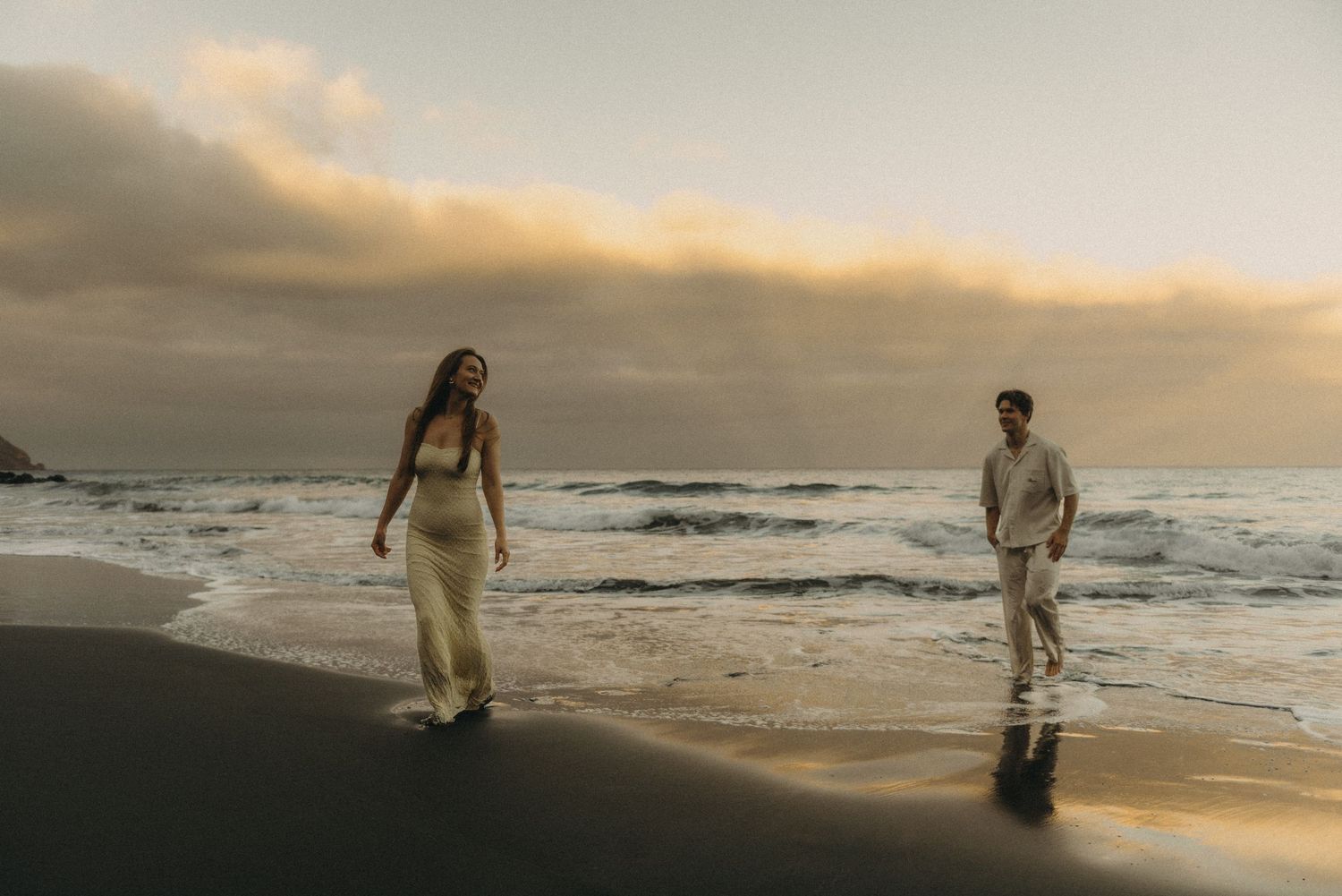 Couple walking on a sandy beach at sunset with golden clouds and ocean waves in the background.
