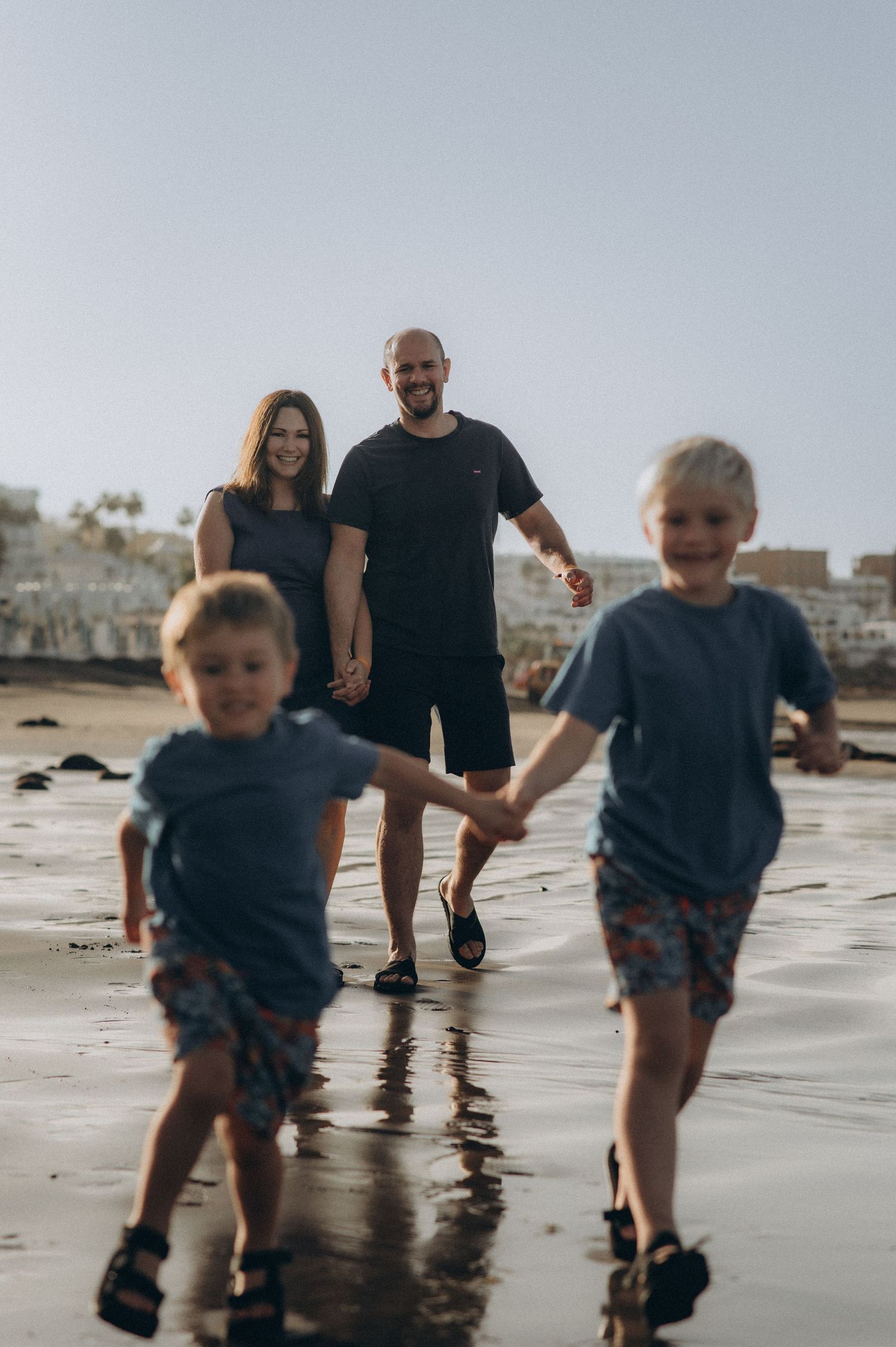 Tender family moment with brothers running together toward the sea while parents observe them in a warm hug at Playa Fañabé