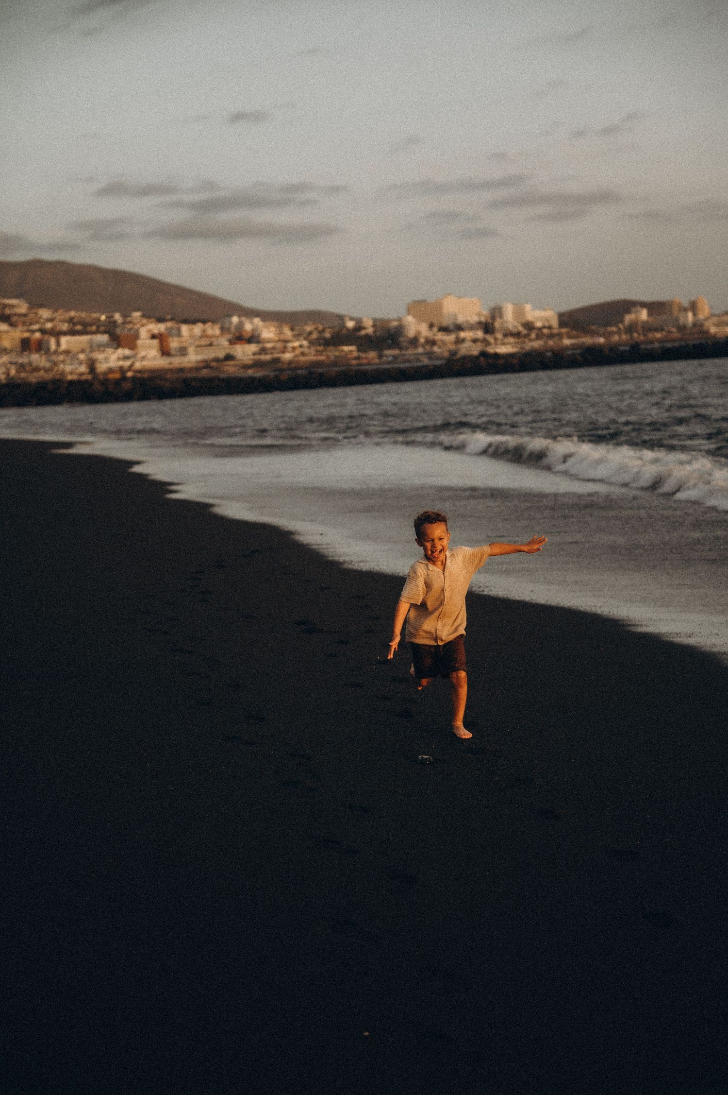Young boy running with arms outstretched along black sand beach by ocean, Playa del Duque