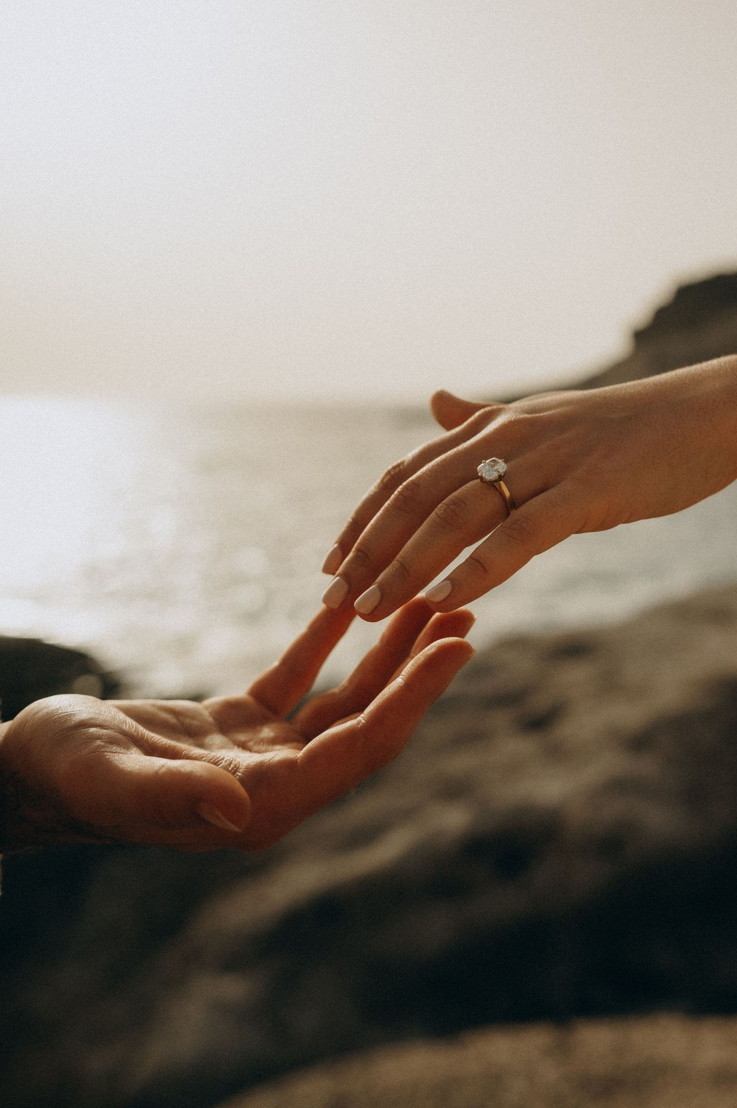 Two hands touching near rocky seaside, one adorned with a stunning diamond engagement ring on gold band.