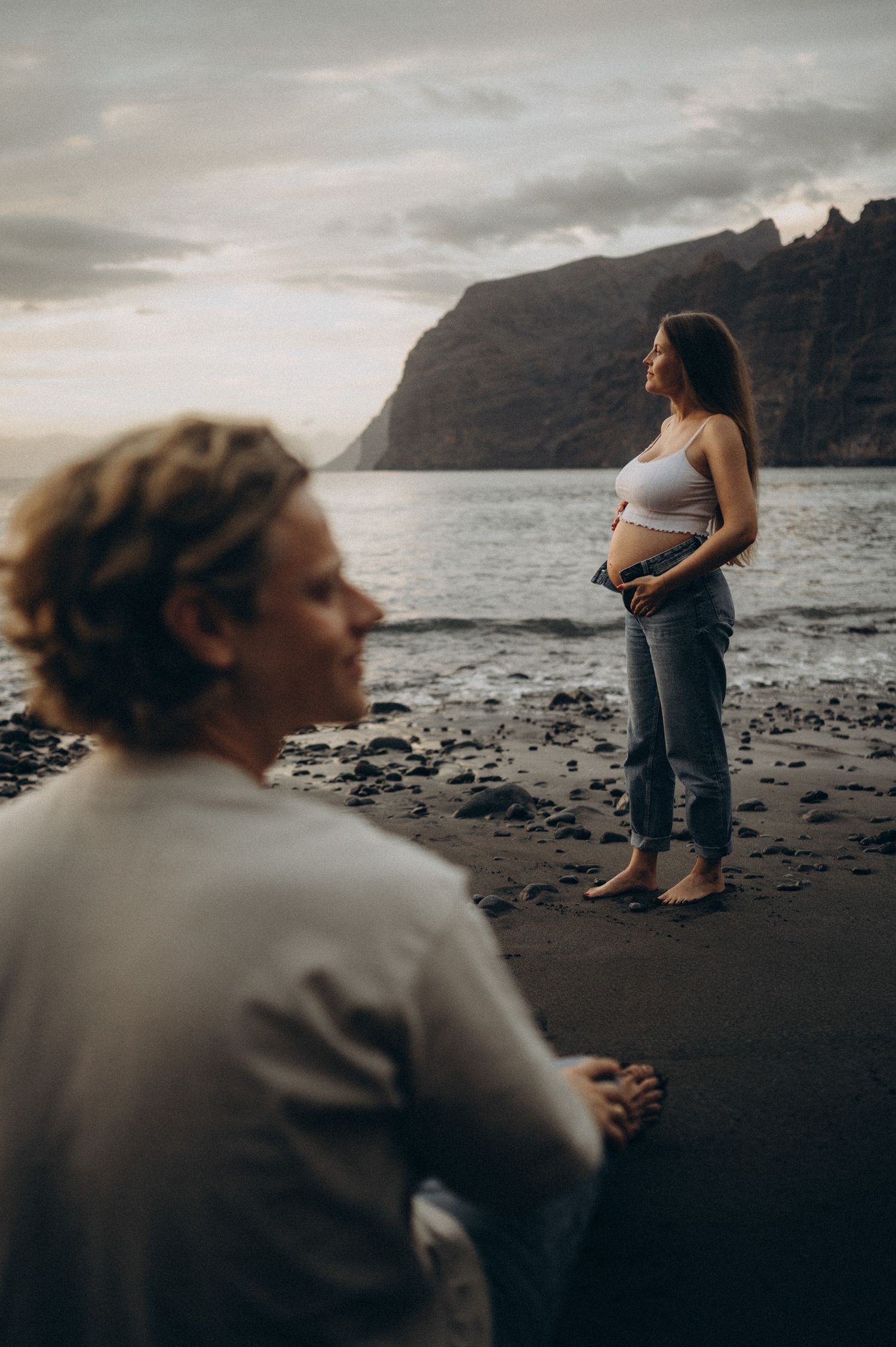 Pregnant woman standing on black sand beach at sunset, cradling her belly, with dramatic cliffs and ocean in background.