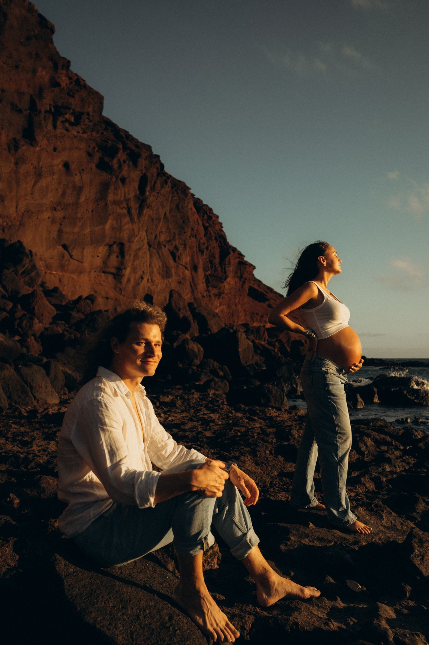 Pregnant couple poses on rocky coastal cliffs at golden hour, man sitting barefoot as woman cradles baby bump.