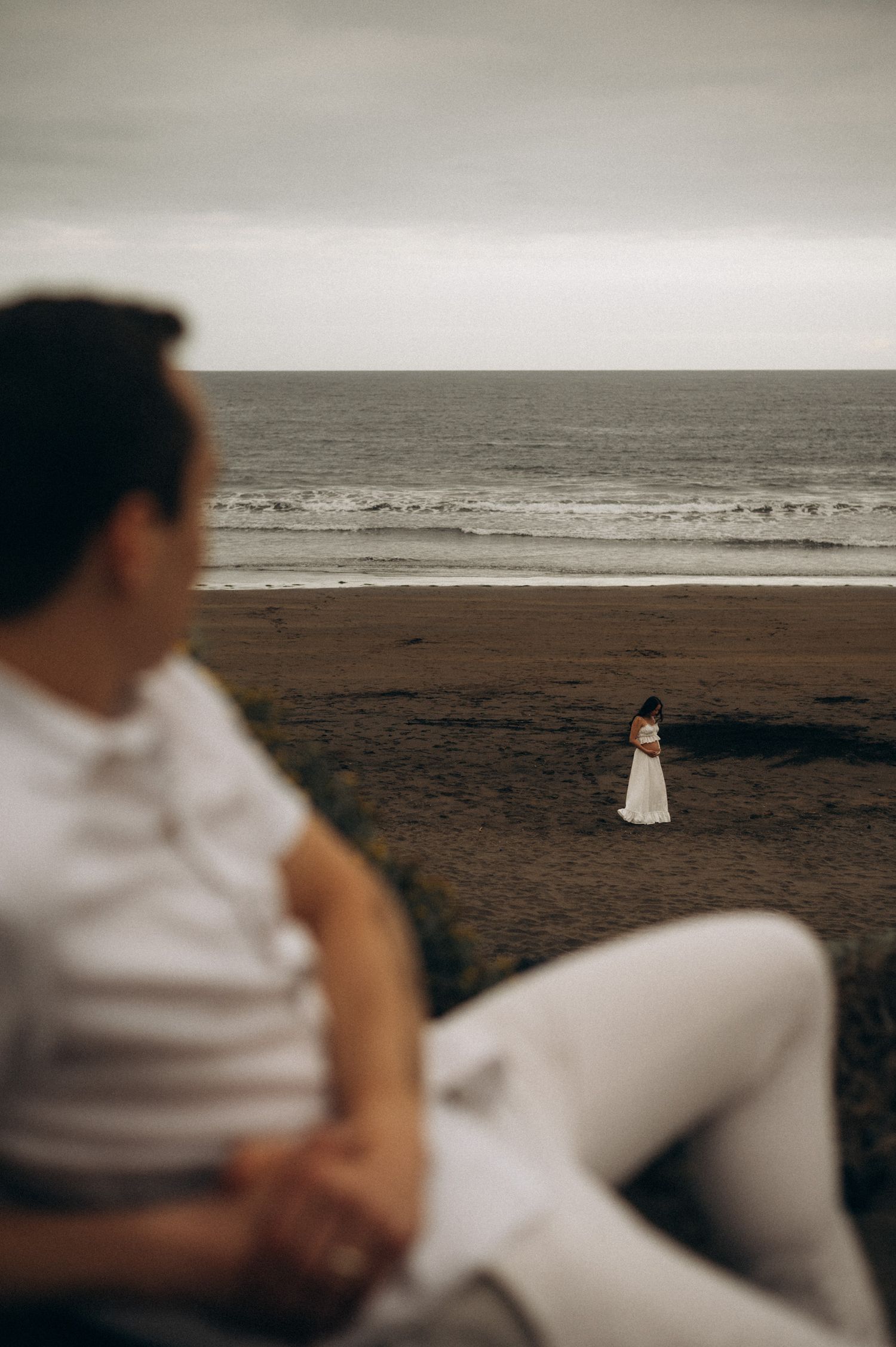 Man in white sitting on cliff watching bride in white dress standing alone on a moody overcast beach.