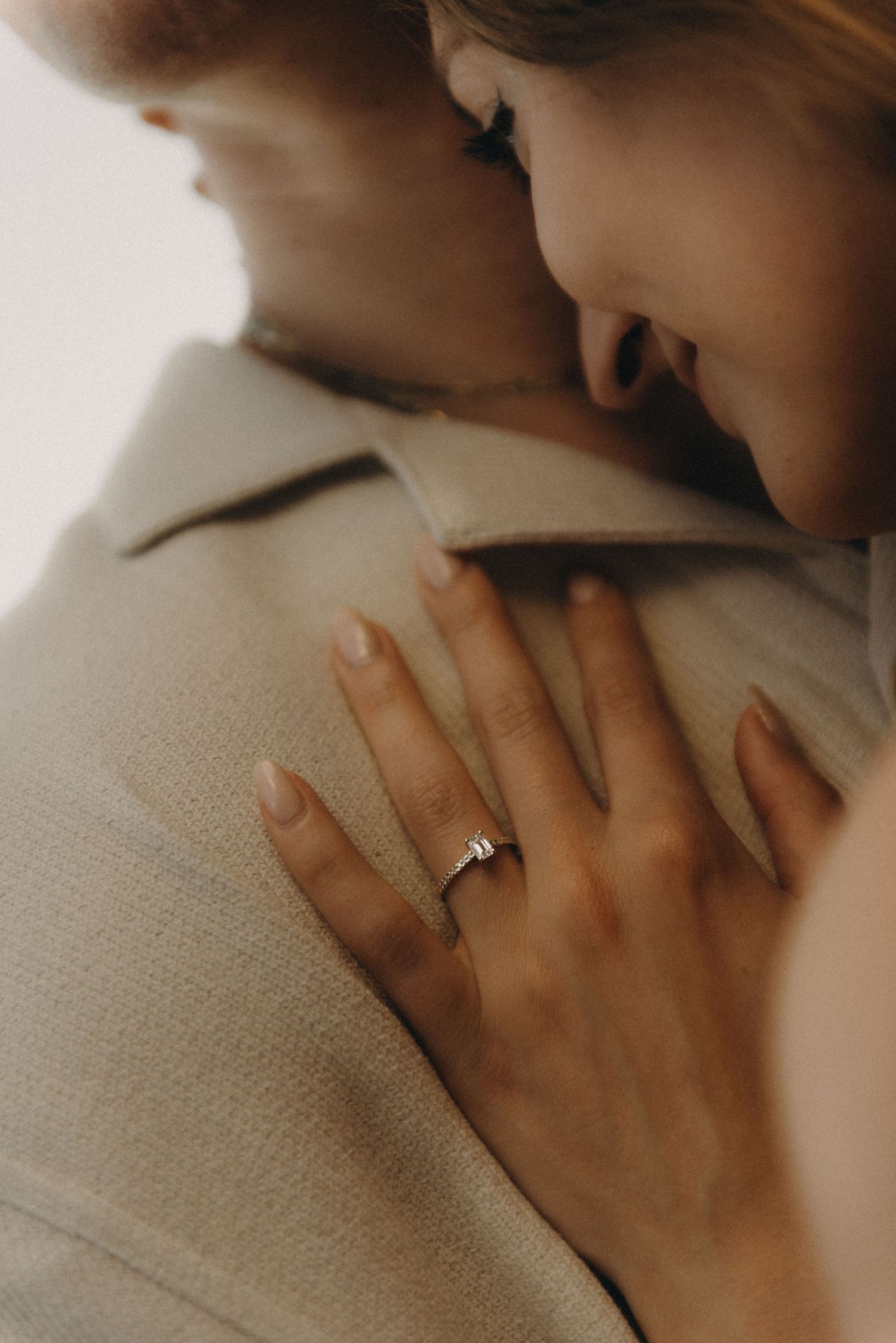 A woman wearing a diamond engagement ring rests her hand on a man's shoulder in a tender embrace.