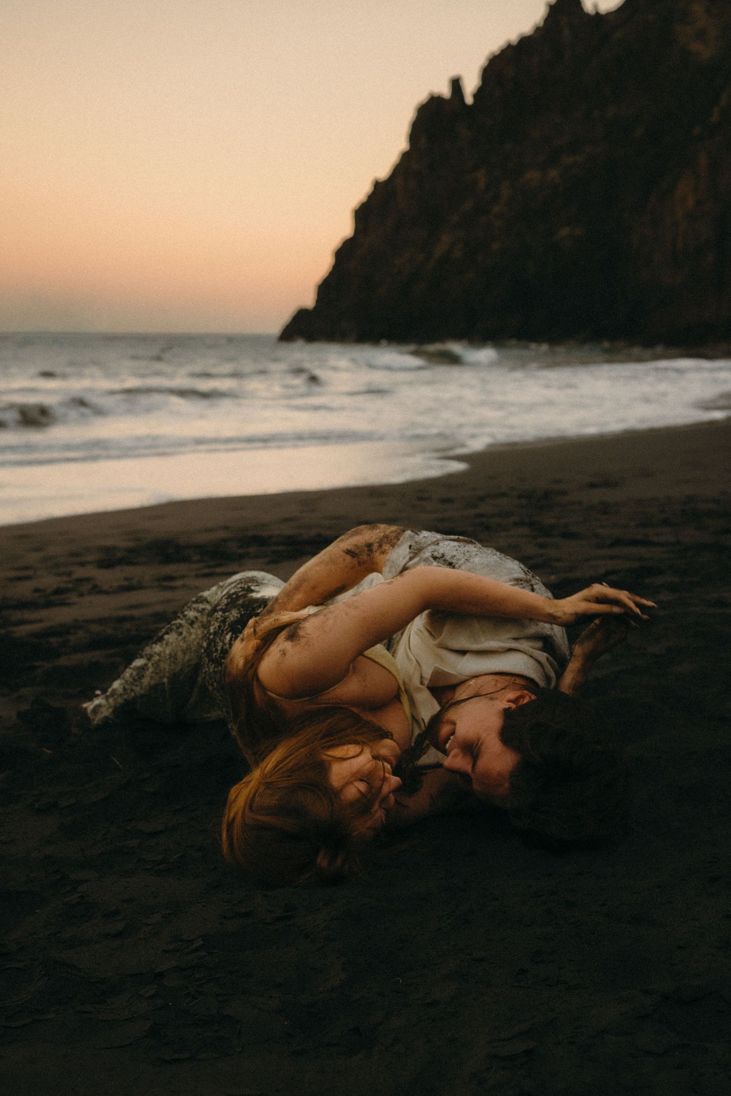Couple lying together on dark sandy beach at sunset with ocean waves and rocky cliffs in background.