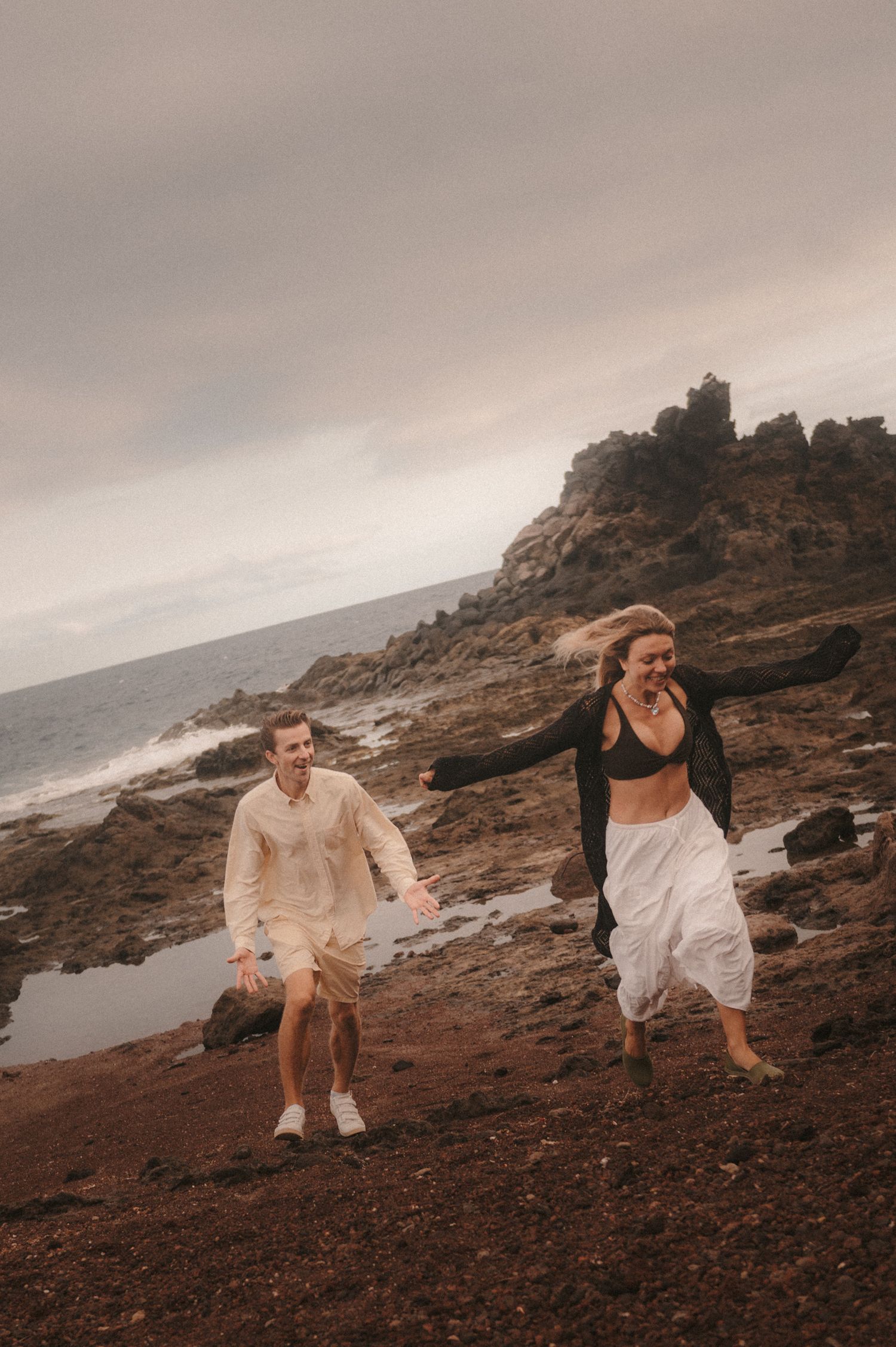 Loving couple playing chase on the rocks by the ocean under Montaña Roja, Tenerife, enjoying a fun and romantic moment