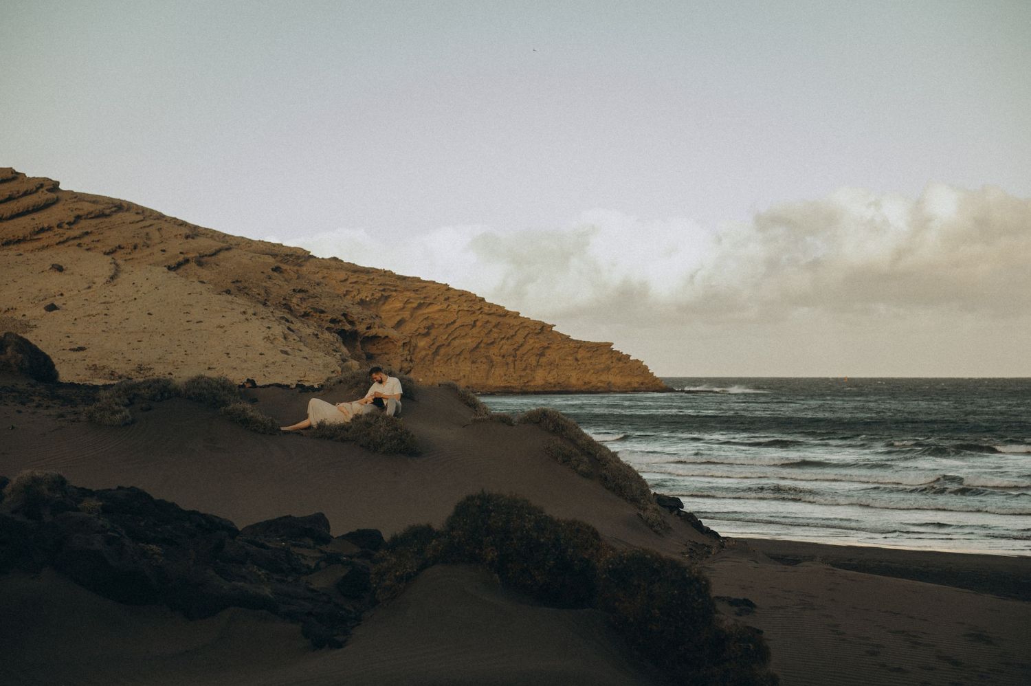 A person in white dress sits on dark volcanic rocks beside a sandy beach with ocean waves and rocky cliffs.
