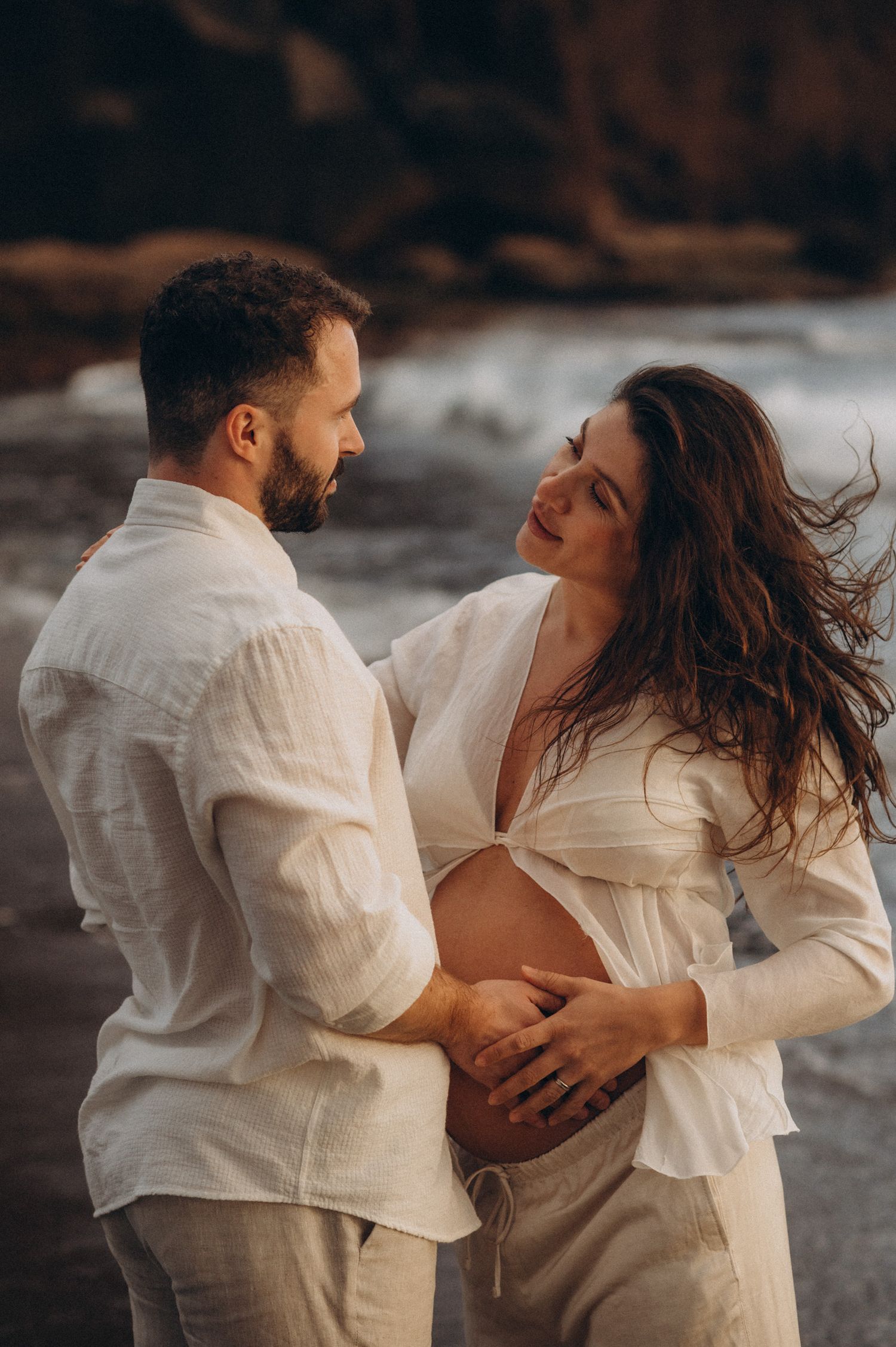 Couple in white linen clothing sharing a tender moment on a rocky beach, woman showing pregnant belly.