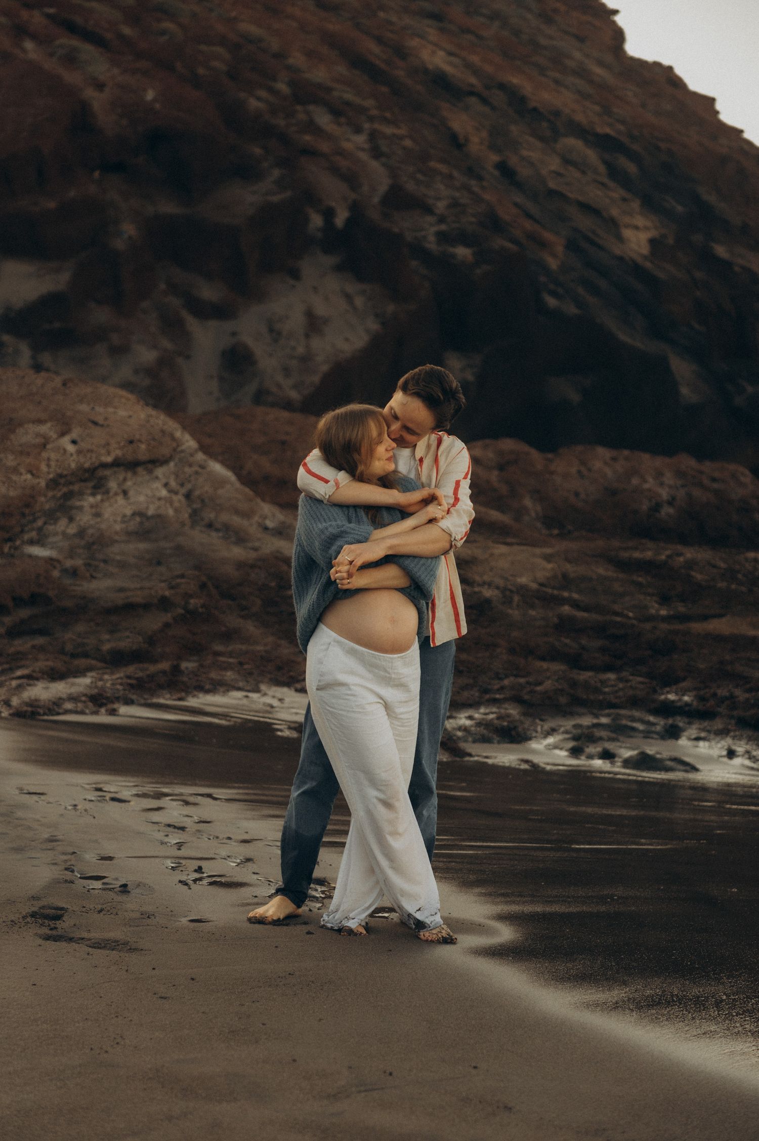 Couple embracing on a dark sand beach, pregnant woman in white pants hugging partner near rocky cliffs.