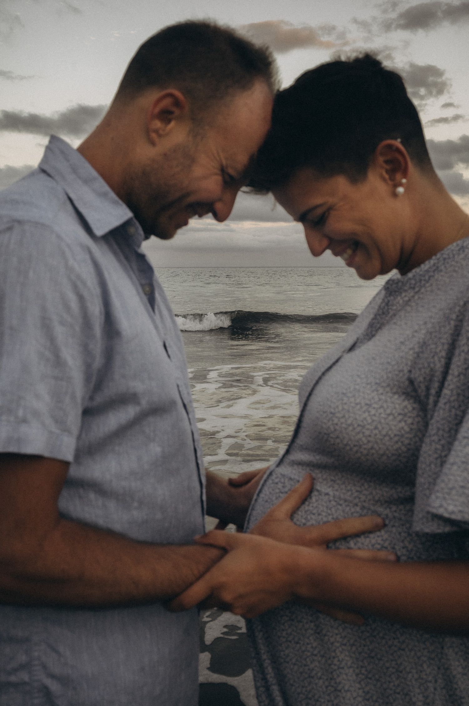 Couple sharing a tender moment on the beach, cradling a pregnant belly, with ocean waves in the background.