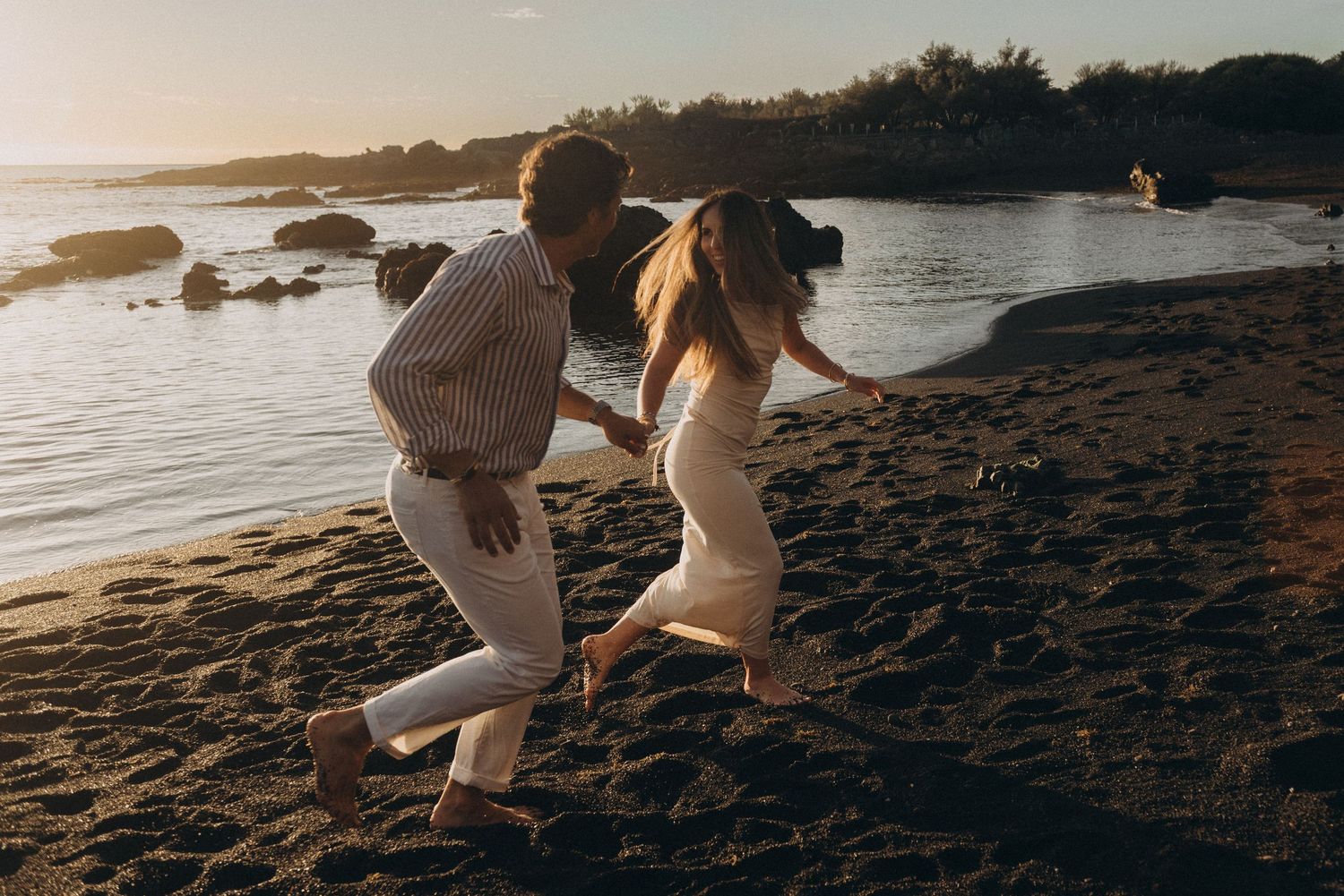 Couple running barefoot on a rocky beach at golden sunset, holding hands in romantic coastal setting.
