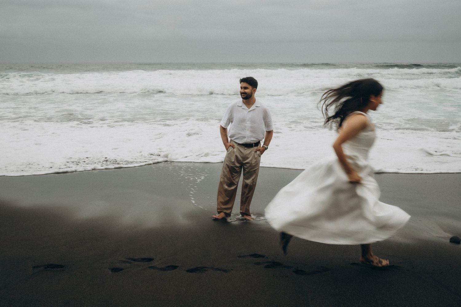 Couple on black sand beach, man in casual attire watches woman in white dress twirl near ocean waves on cloudy day.