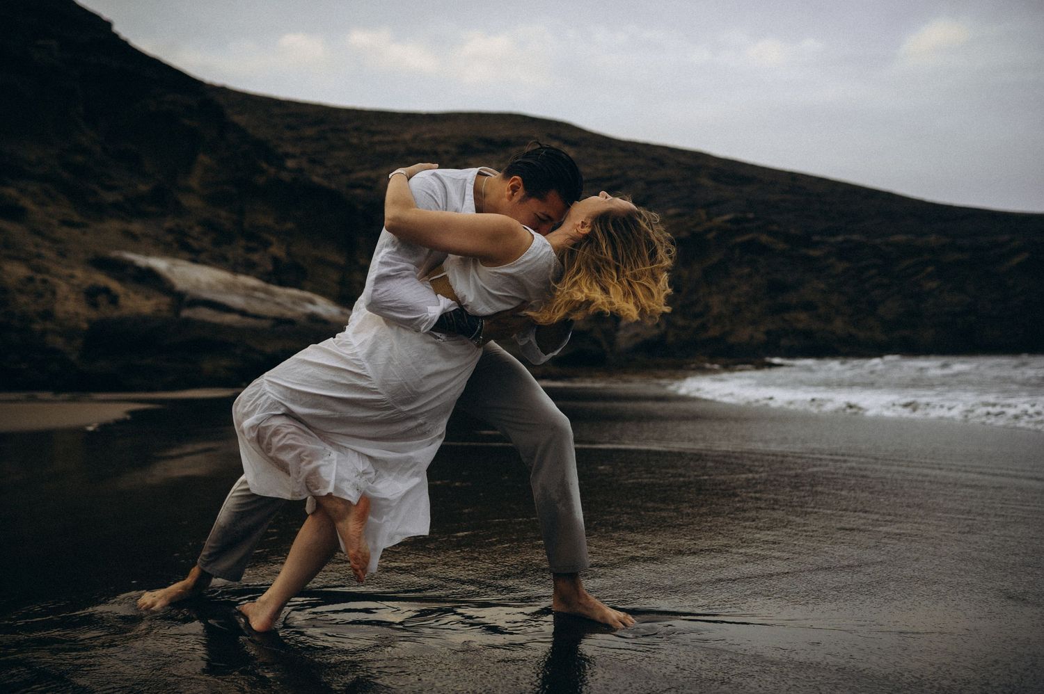 Couple sharing a passionate kiss on a dramatic dark beach with ocean waves and rocky cliffs in background.