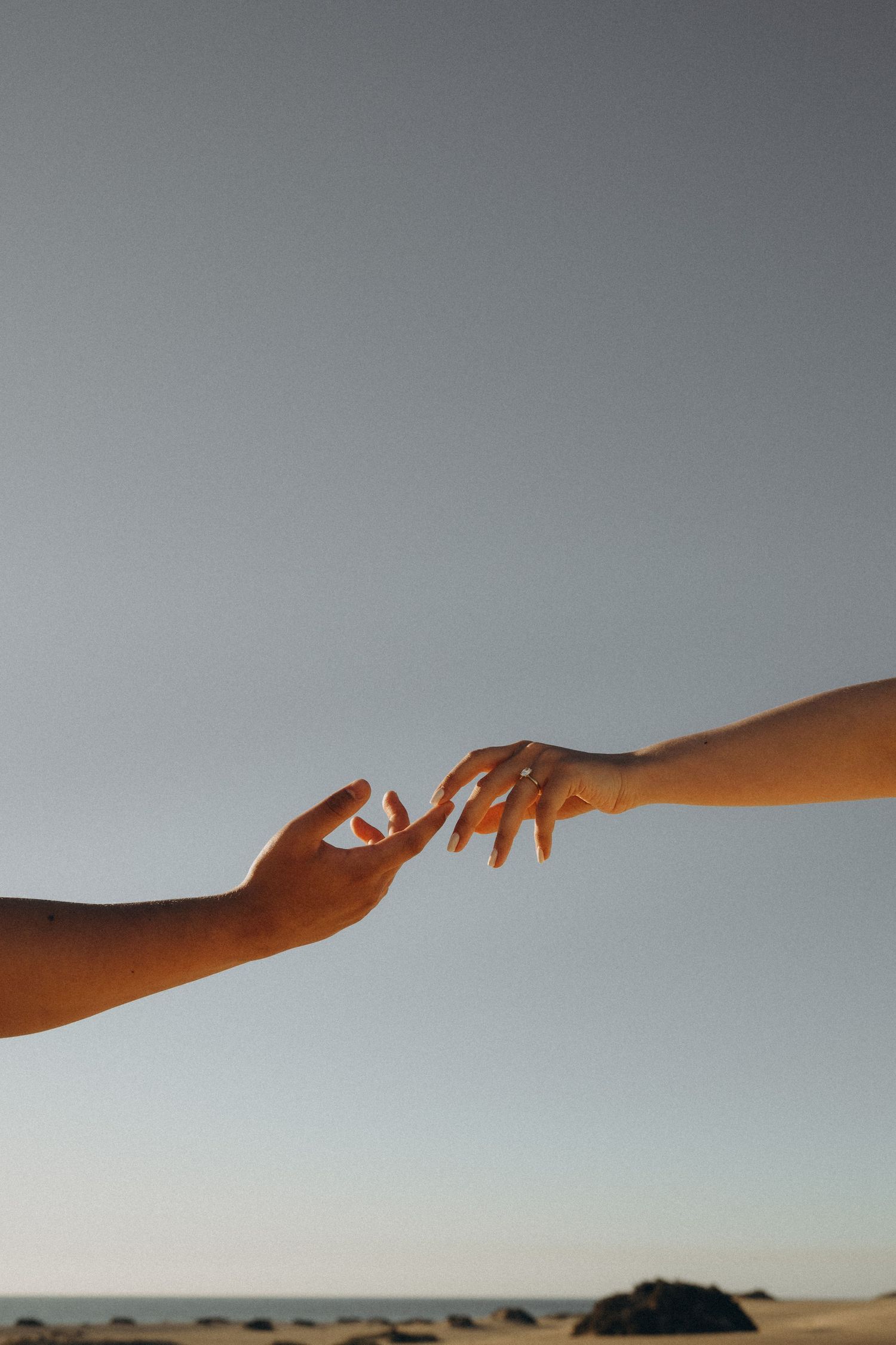 Two hands reaching toward each other against a clear sky at dusk near a beach.