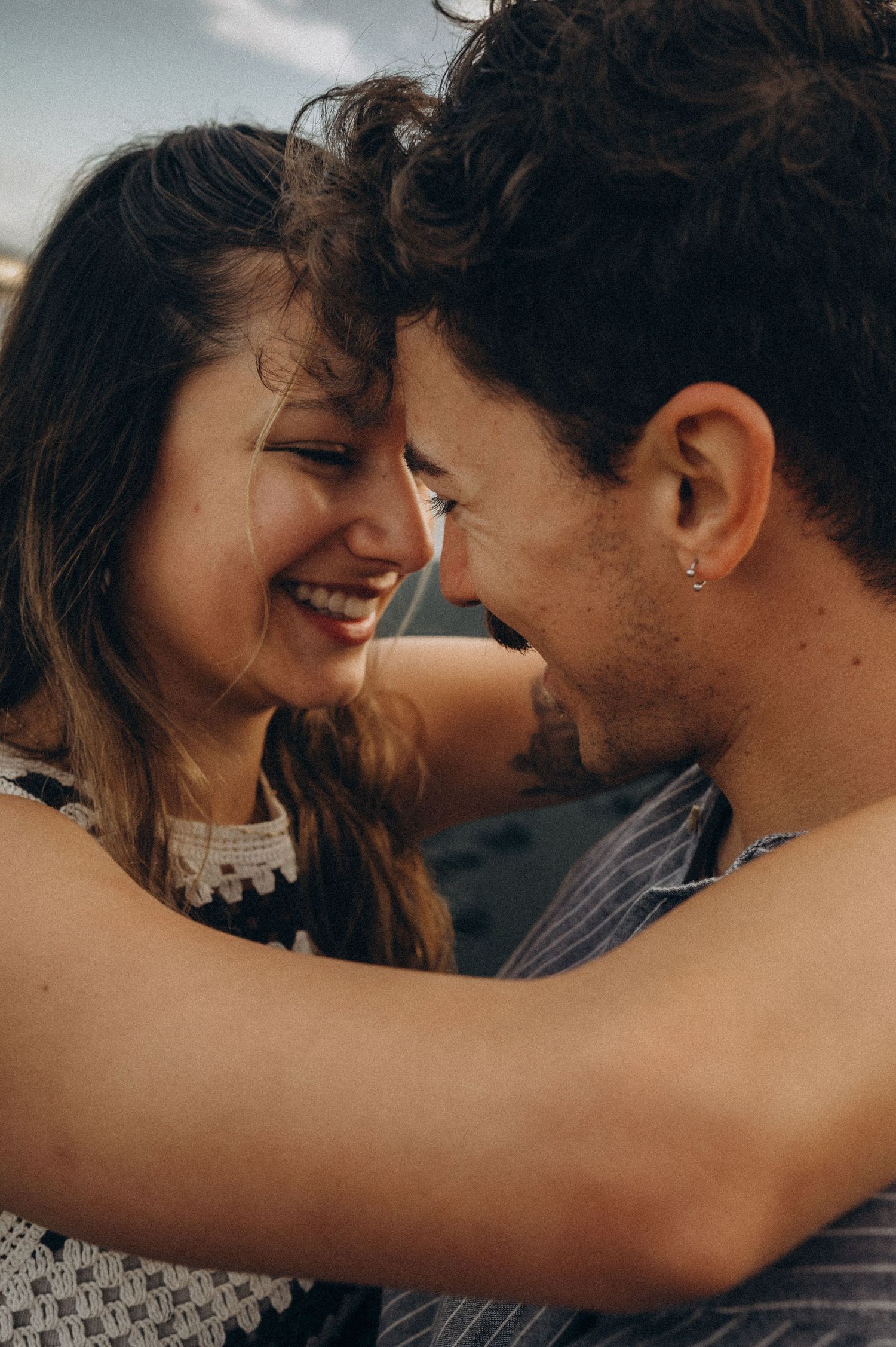 A smiling couple sharing a romantic moment, foreheads touching, embracing outdoors with soft natural lighting.
