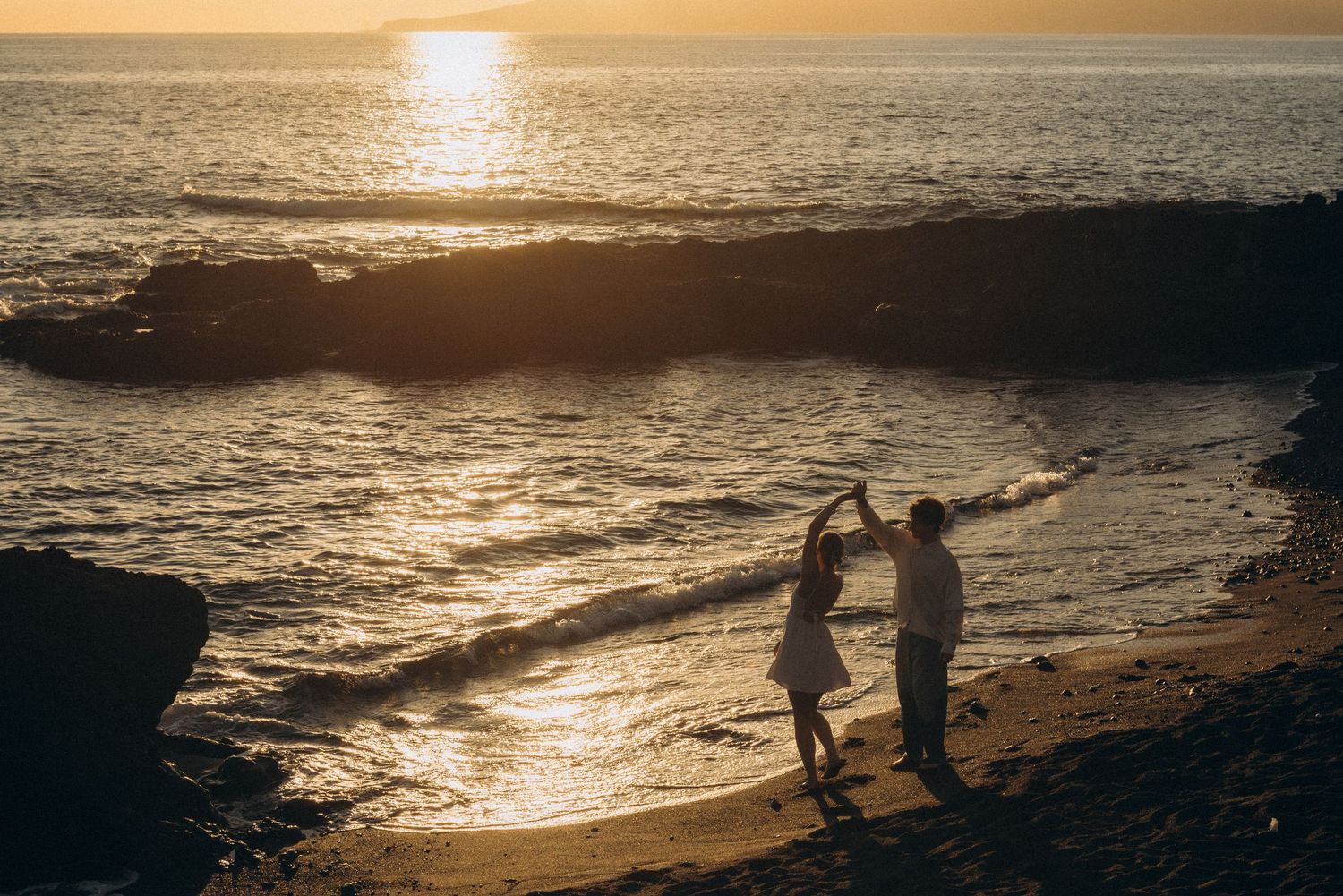 Couple dancing romantically on a sandy beach at golden sunset with ocean waves and rocky coastline in background.