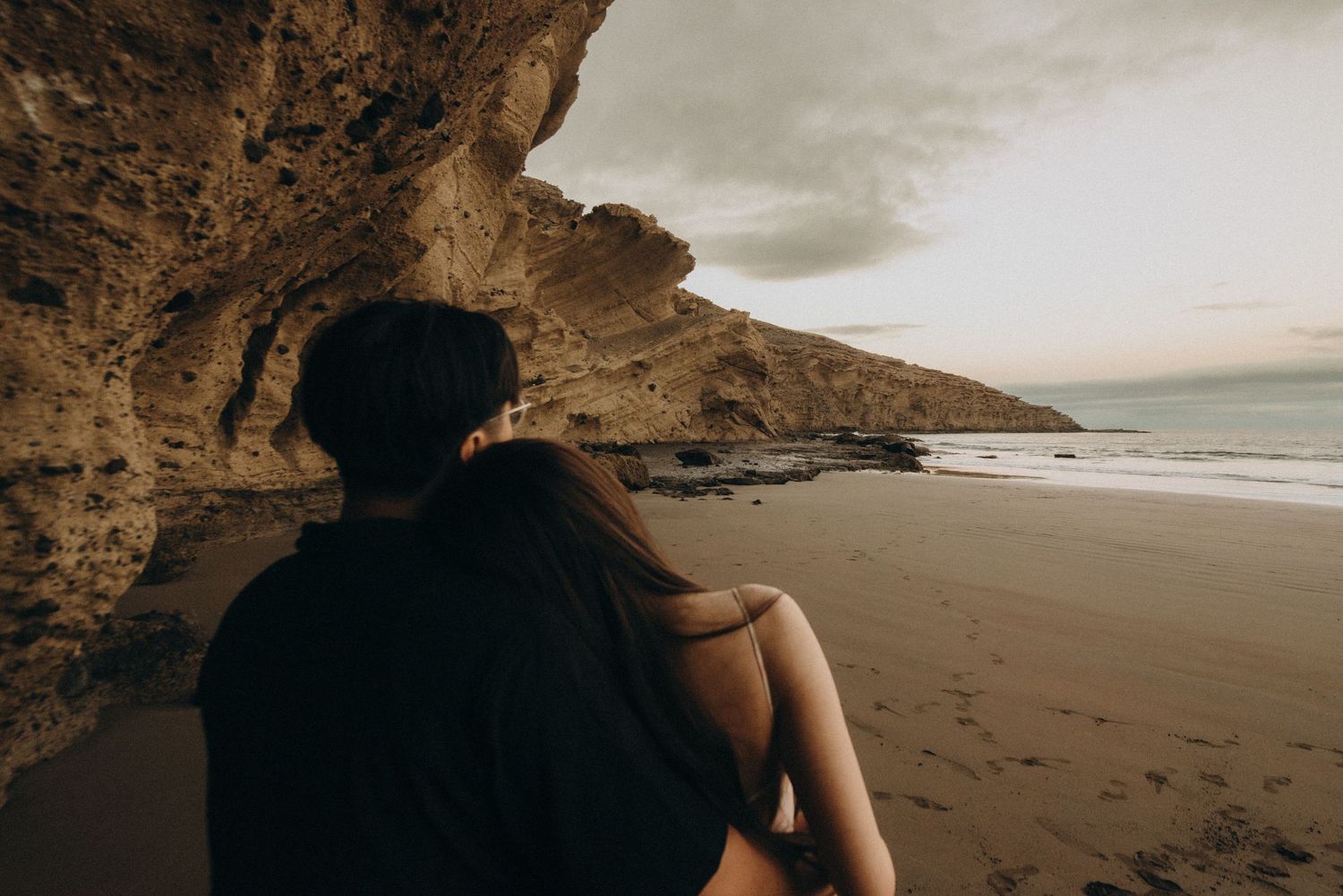 Couple sitting together under rocky coastal cave overlooking sandy beach with dramatic cloudy sky at sunset.