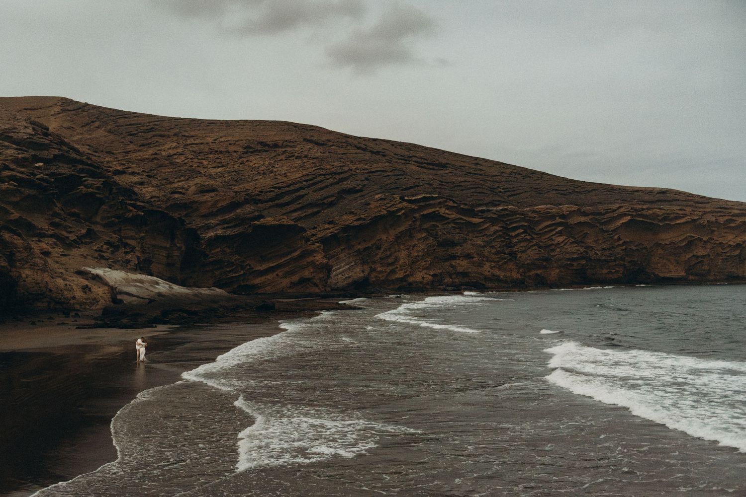 A lone figure walks along a dramatic black sand beach with dark volcanic hills and moody ocean waves under overcast skies.