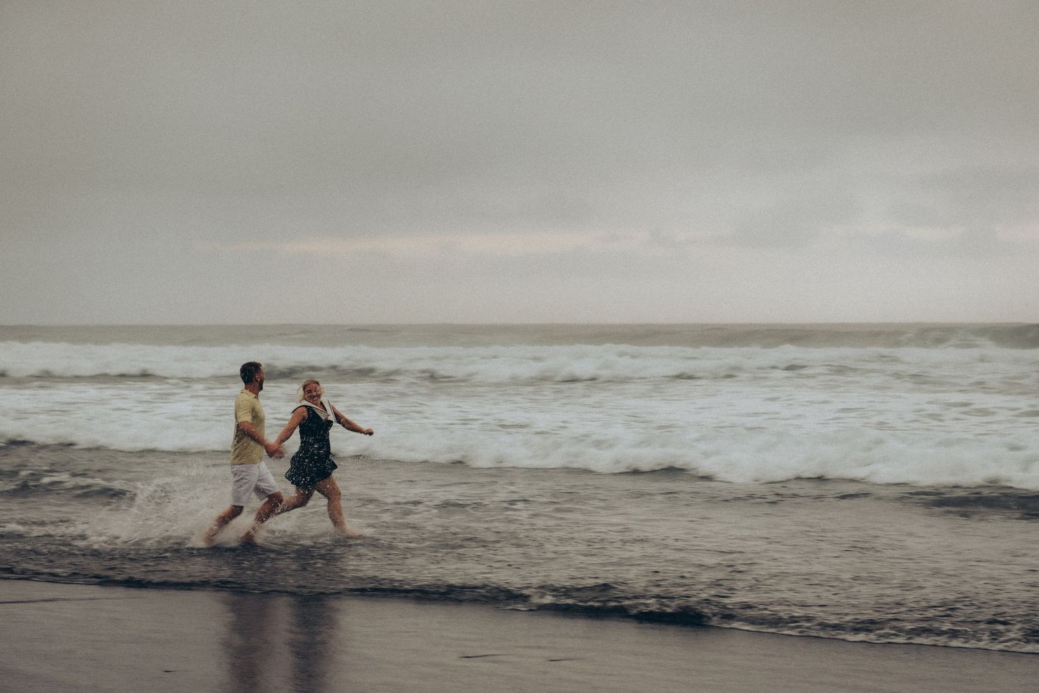 Couple holding hands running along a sandy beach shore with ocean waves on a cloudy day.