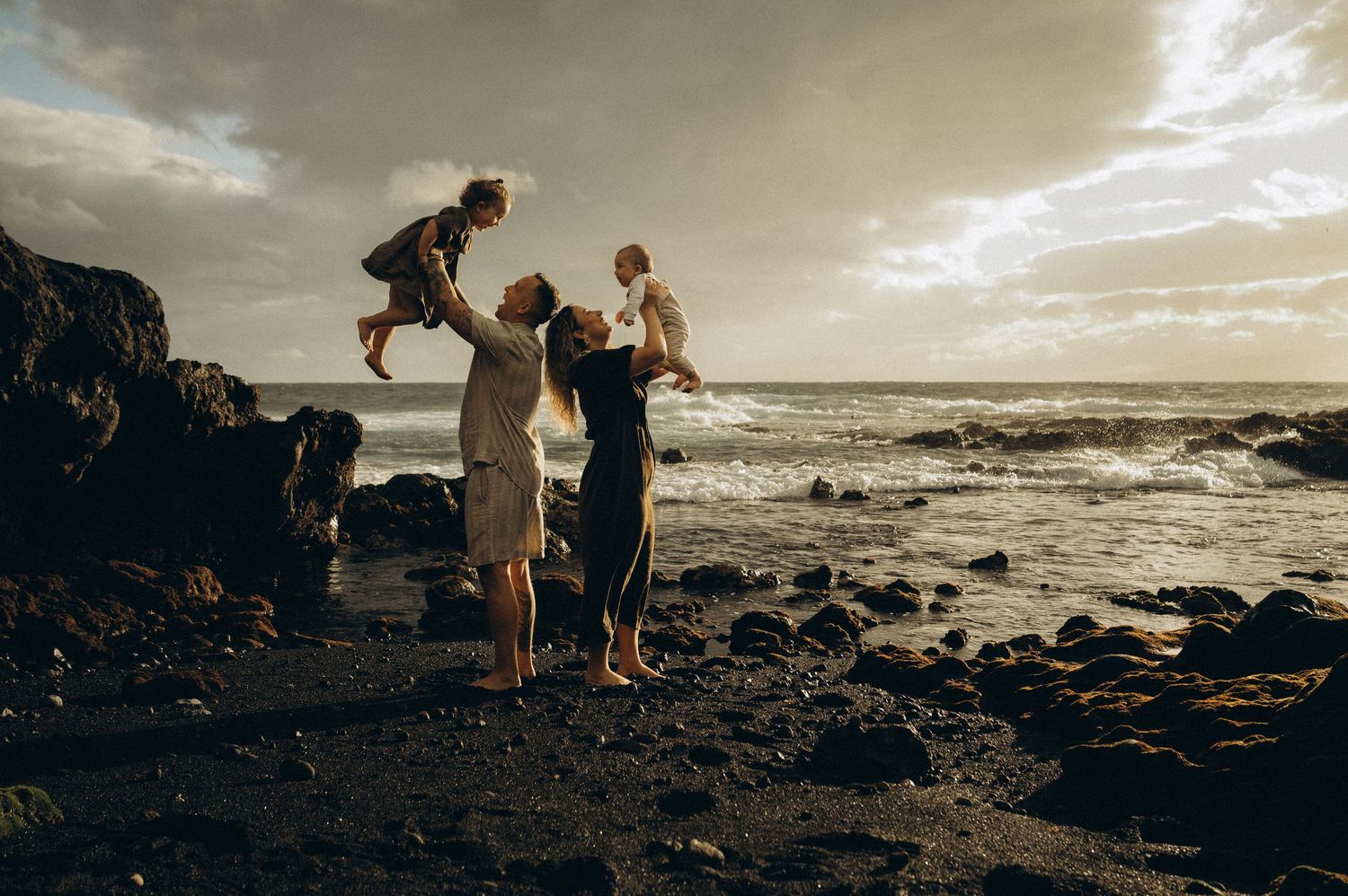 Family playing on rocky beach at sunset, parents lifting children in the air with ocean waves in background. Playa La Jaquita, Tenerife