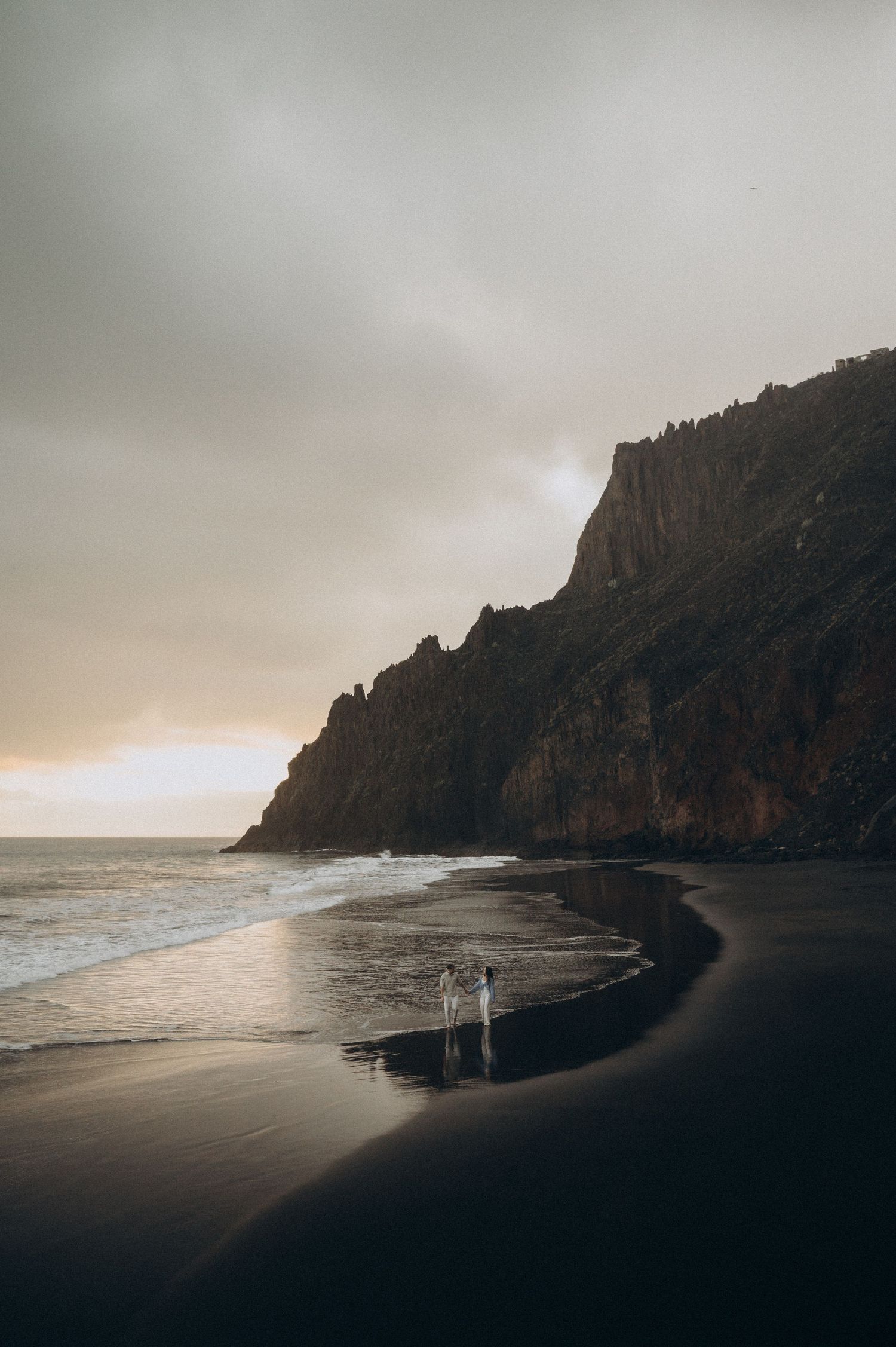A lone figure stands on a dramatic black sand beach at sunset with towering cliffs and crashing waves in Playa Benijo, Tenerife