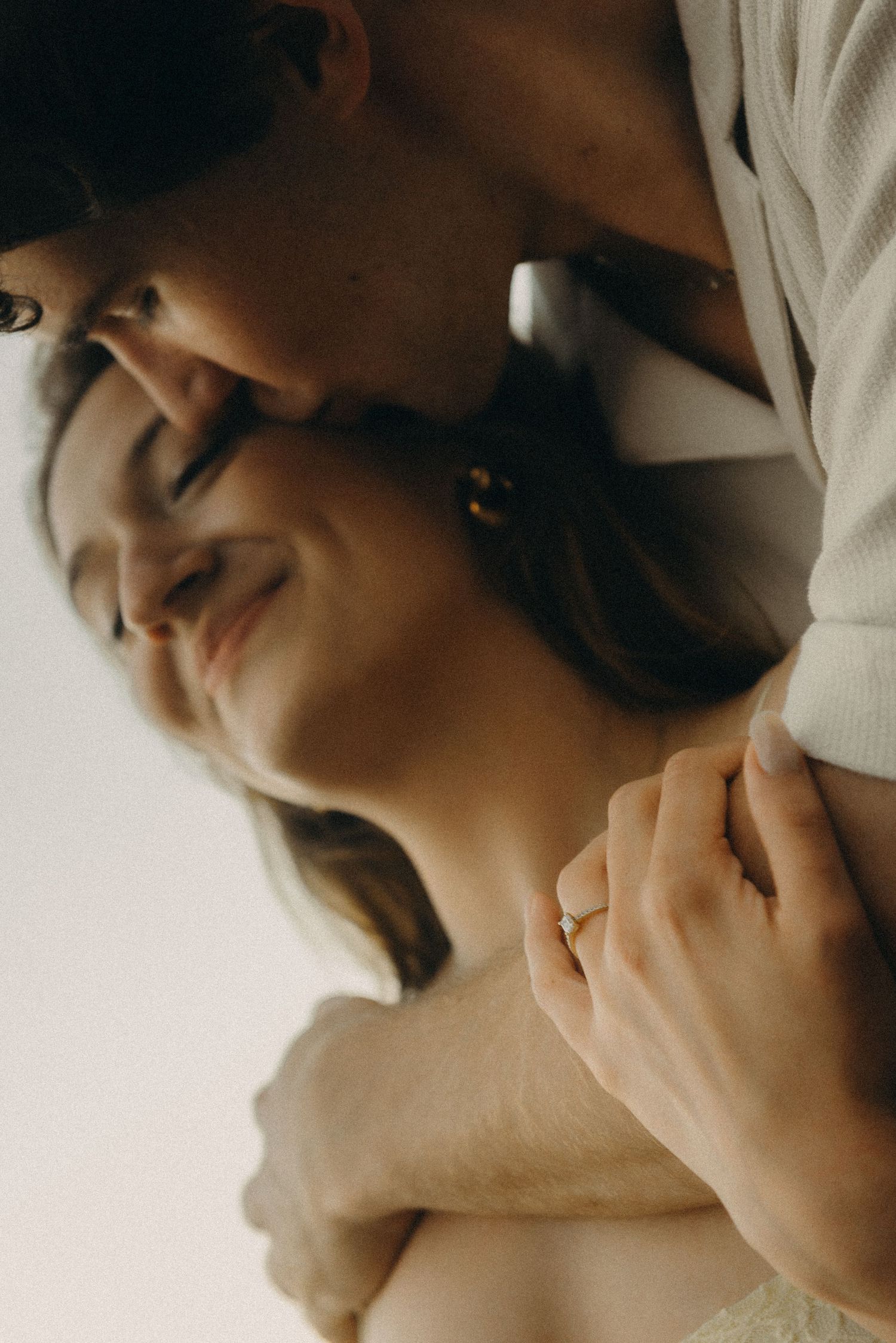 A couple shares an intimate embrace, the groom kissing the bride's head as she smiles softly, wearing a delicate ring.