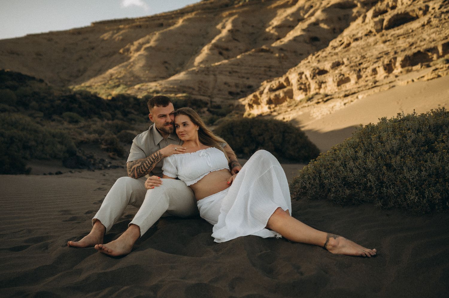 Couple sitting together on sandy desert dunes at golden hour, woman in white dress, man in linen outfit, rocky cliffs behind.