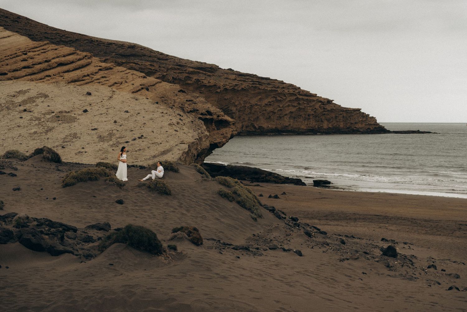 Couple in white dress sitting on volcanic black sand beach with dramatic rocky cliffs and ocean backdrop.