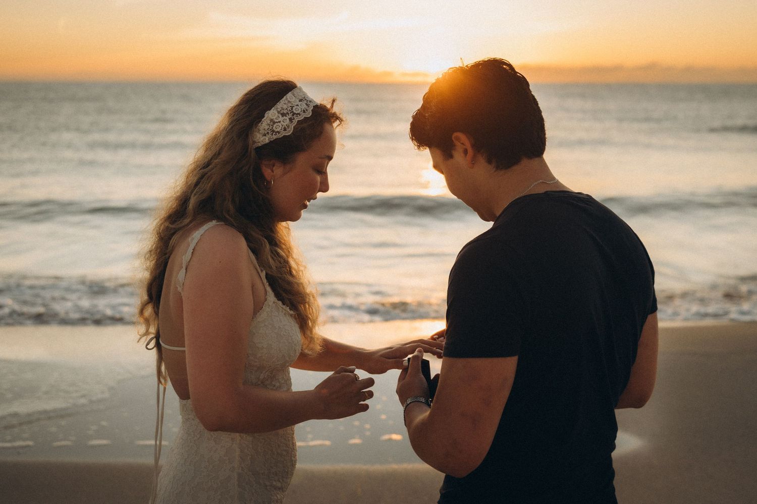 Couple exchanging rings on a beach at sunset, bride in white dress with floral headpiece, groom in black t-shirt.