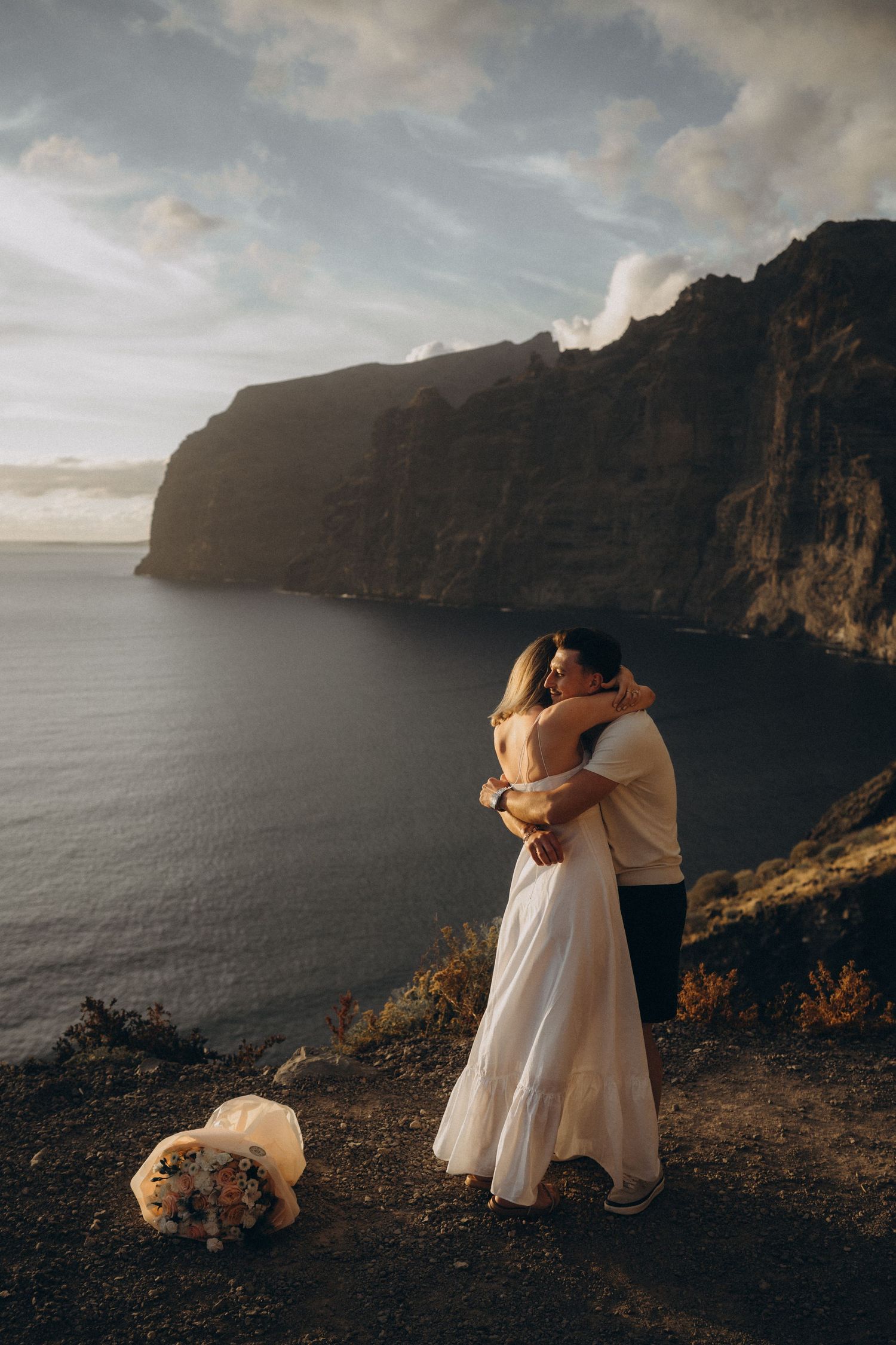 Bride and groom embracing on a cliff overlooking the ocean with dramatic rocky mountains at sunset.