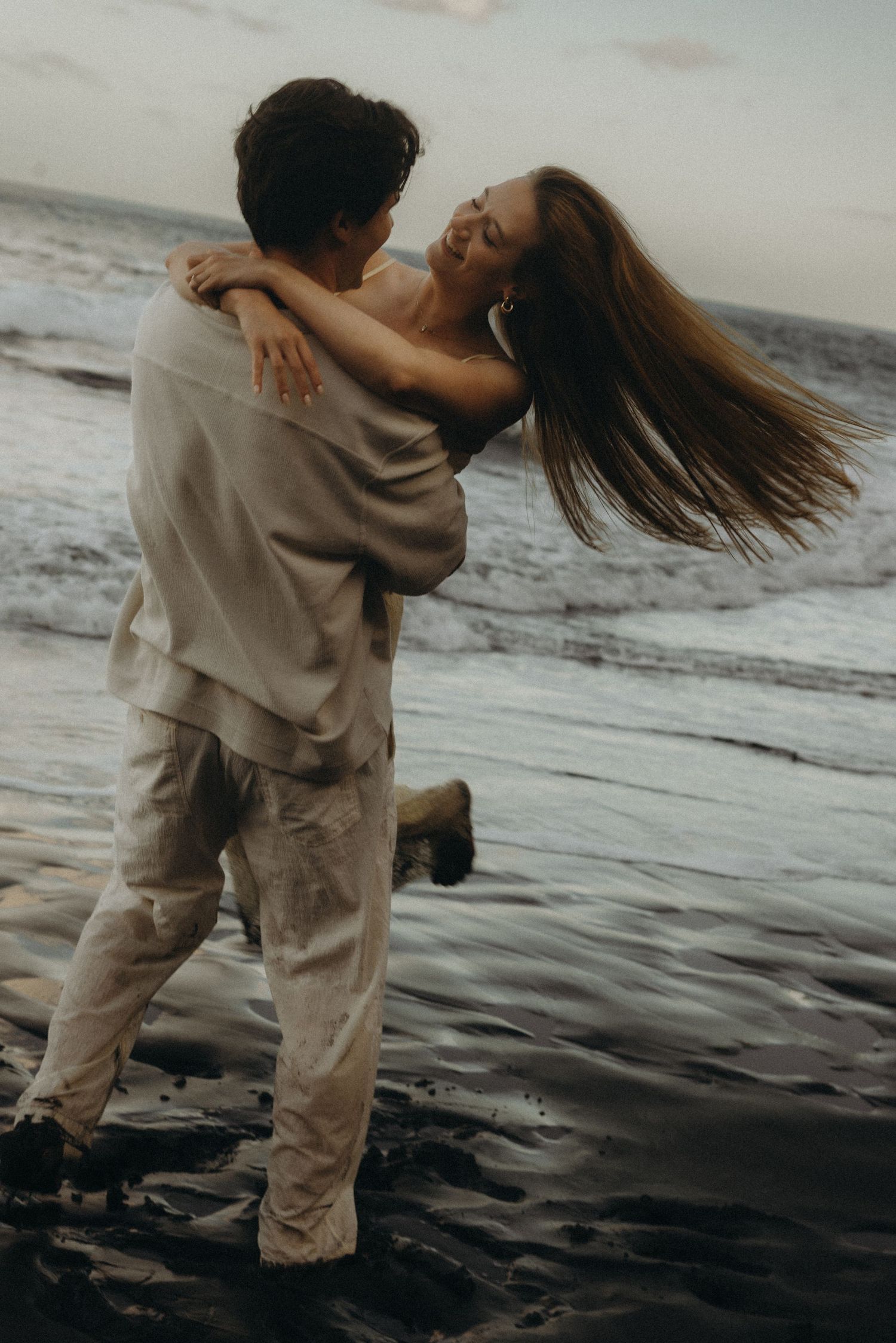 A couple embraces joyfully on a dark sandy beach at dusk, with ocean waves in the background.