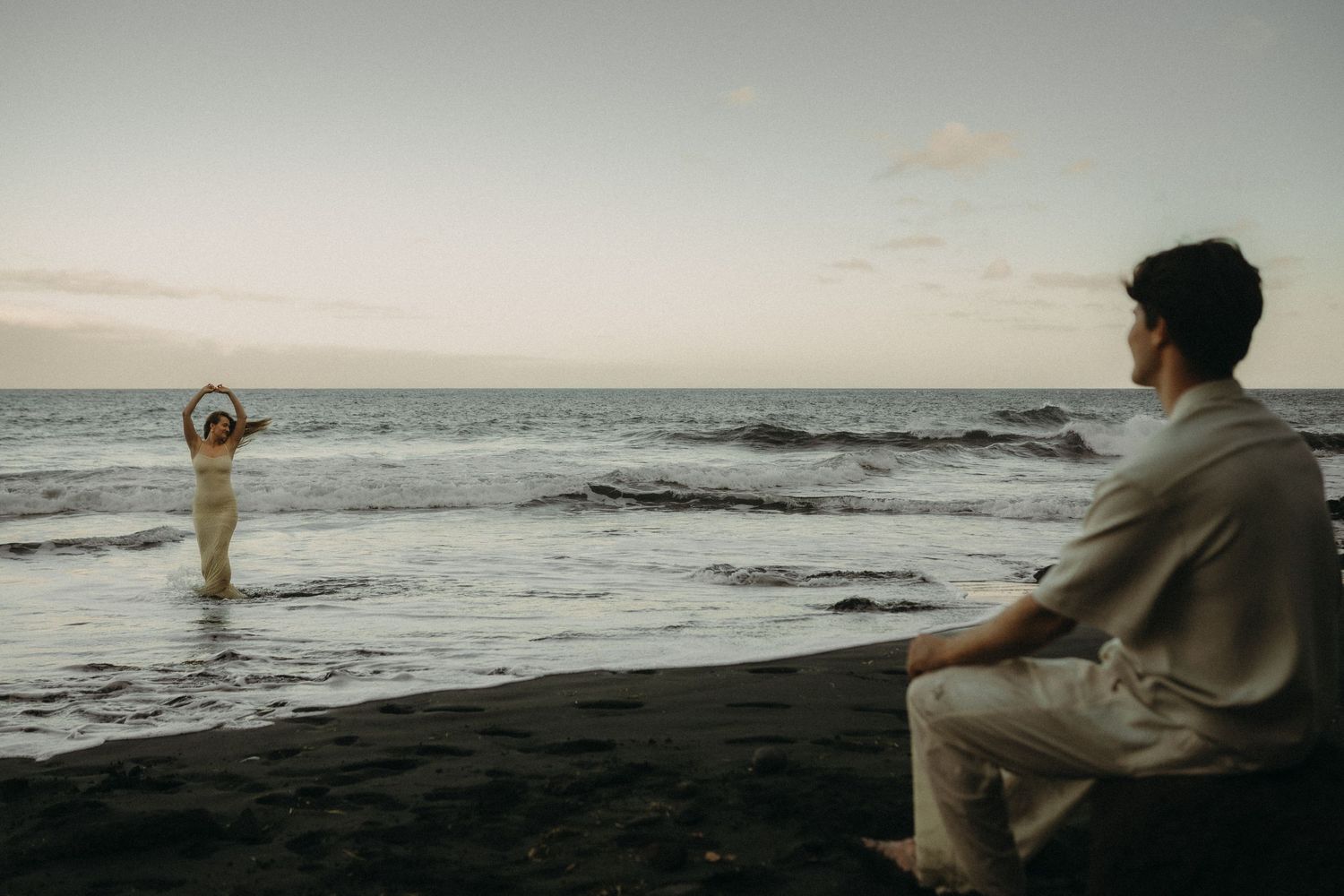 A couple shares a romantic beach moment at sunset, one standing in the waves while the other sits on the shore.