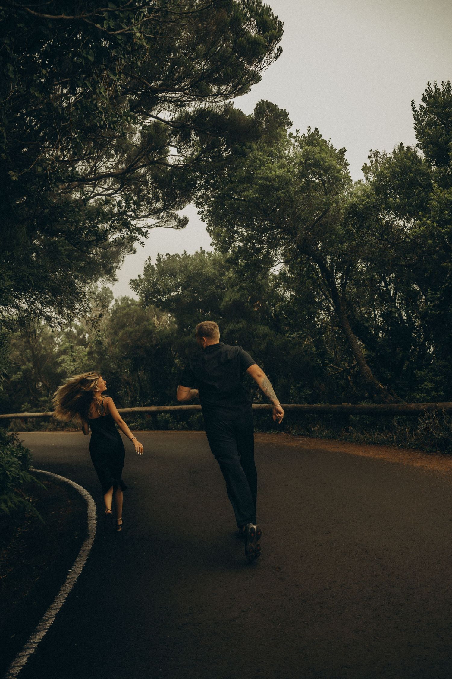 Couple running together on a winding road surrounded by lush green trees in a moody outdoor setting.