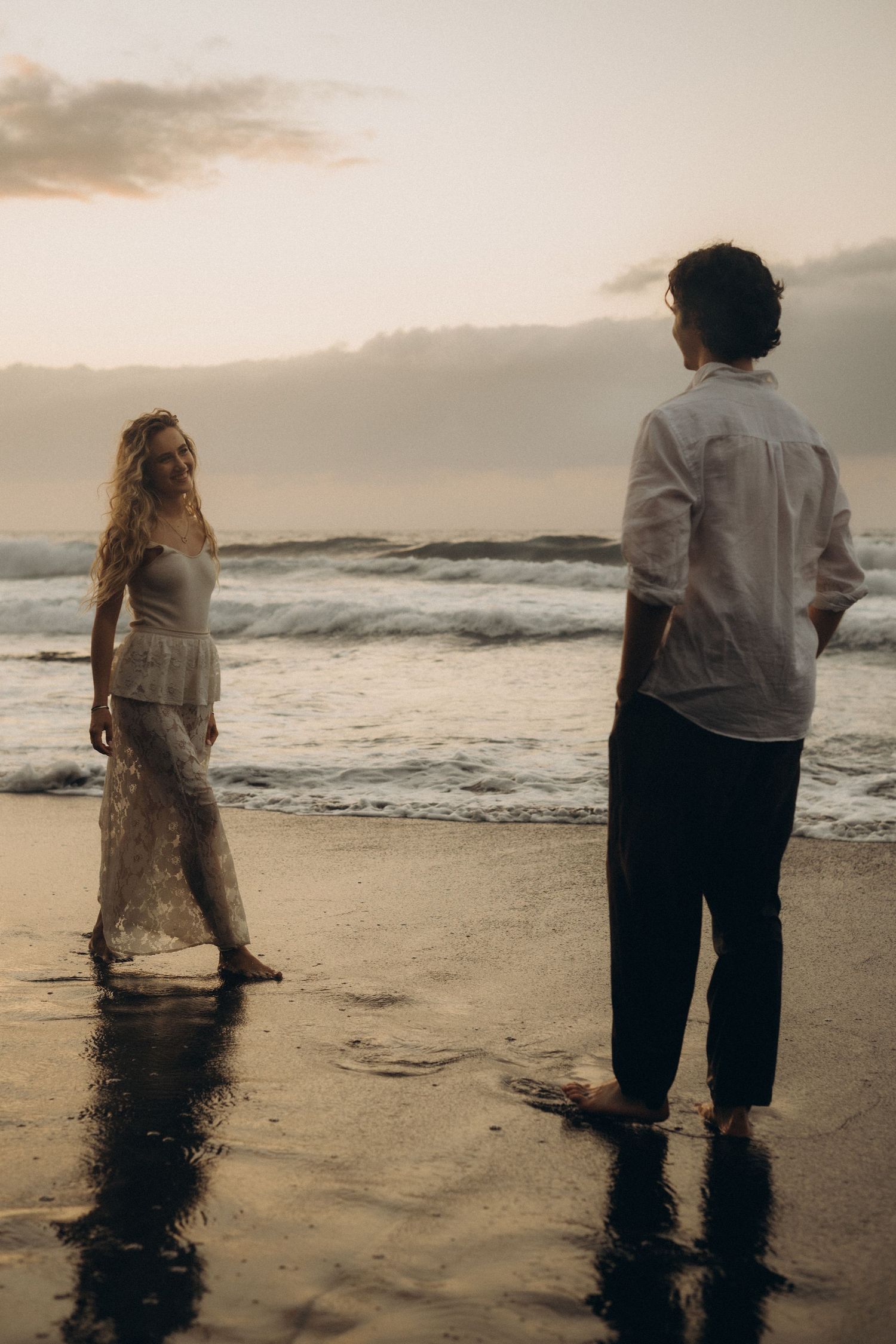 Couple standing barefoot on a wet beach at sunset, woman in white lace dress smiling at man in white shirt.