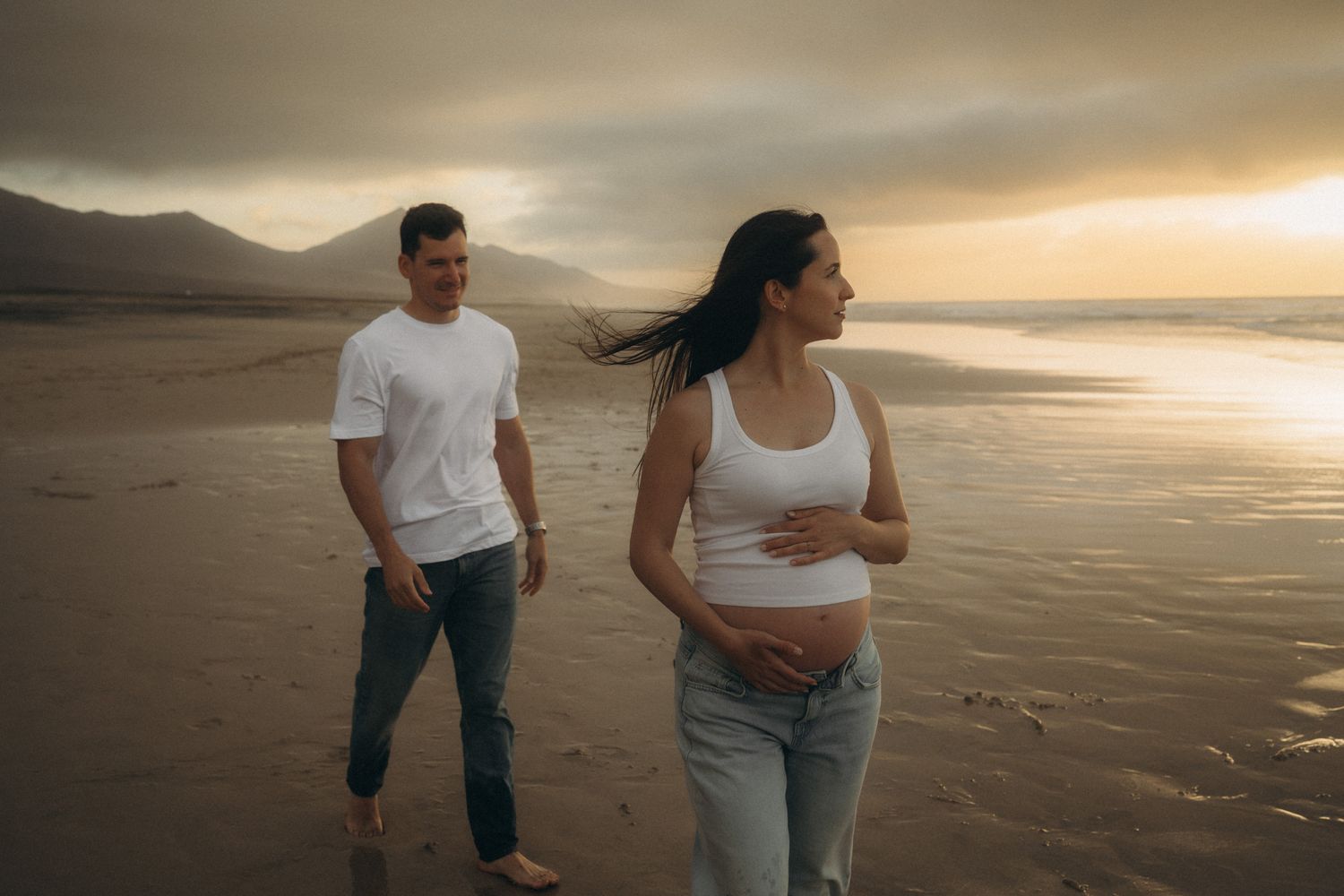Pregnant woman holding belly with partner walking on beach at sunset with mountains in background.