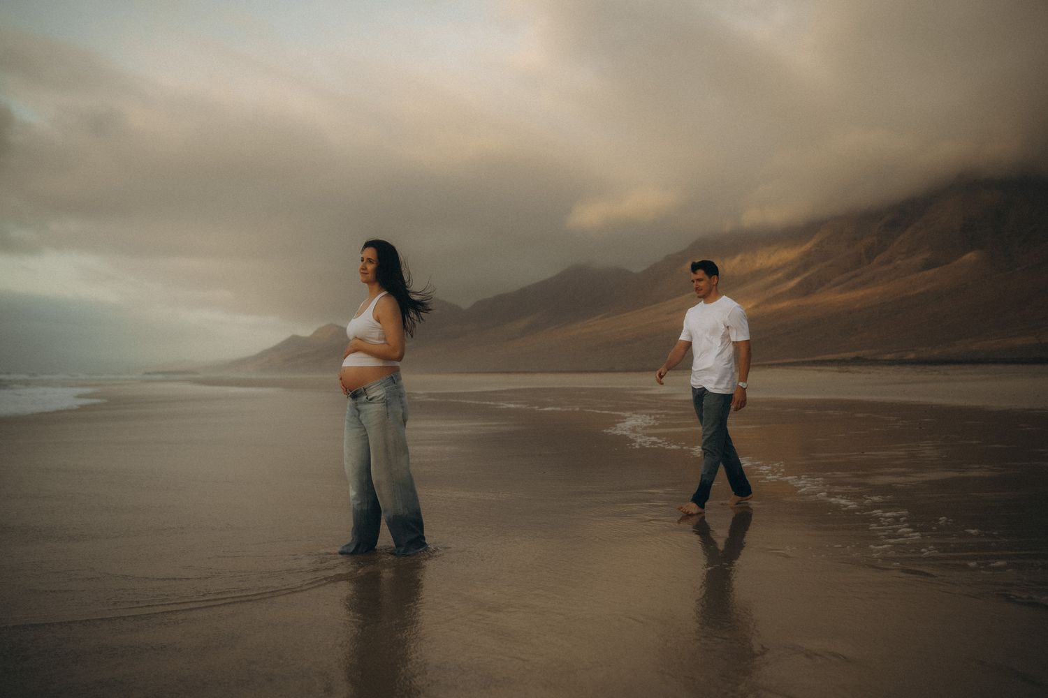 Pregnant woman and man walking on a misty beach with dramatic mountains and cloudy sky in the background.