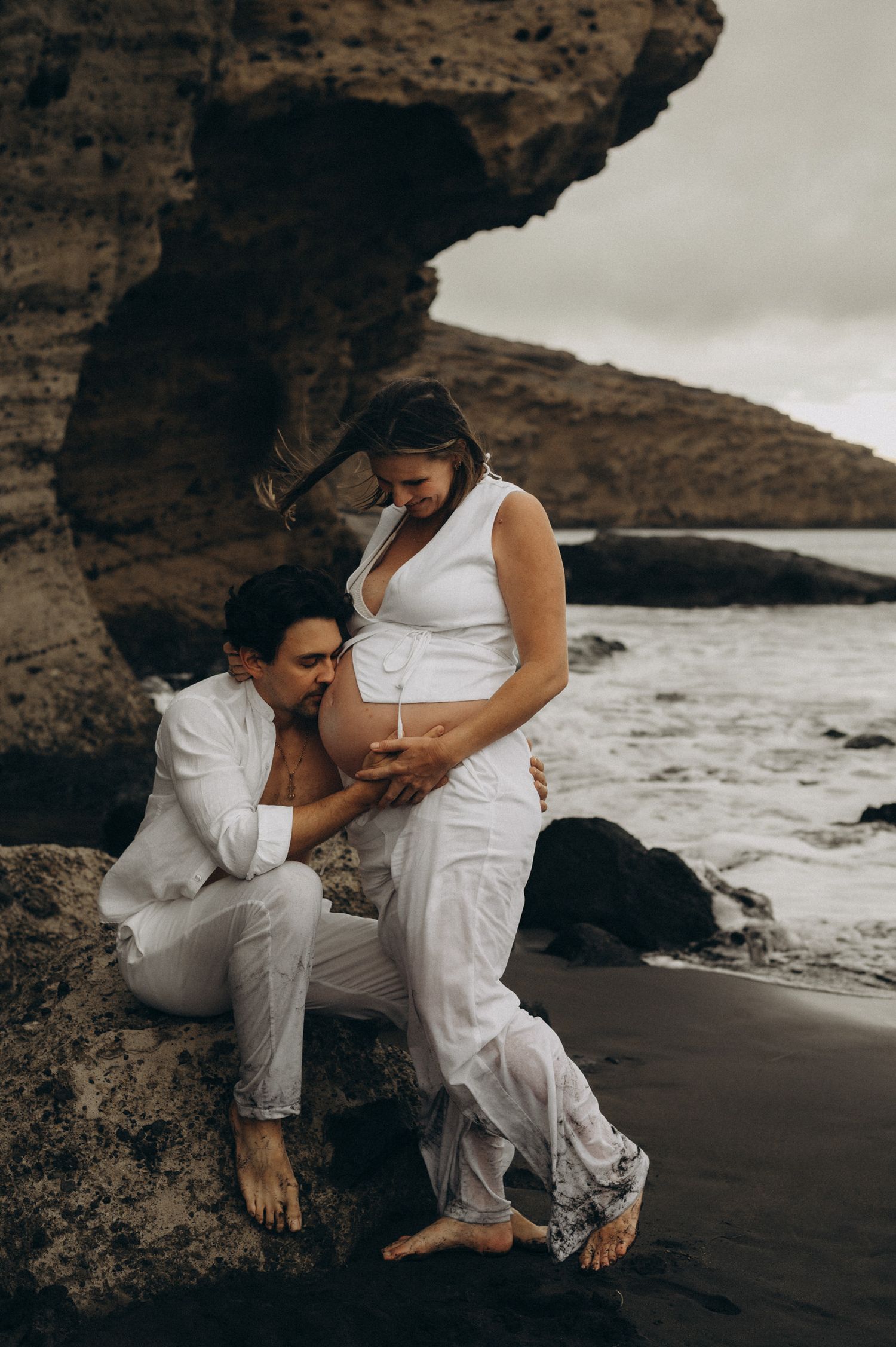 Couple in white outfits on rocky beach, pregnant woman standing while partner kisses her baby bump near ocean.