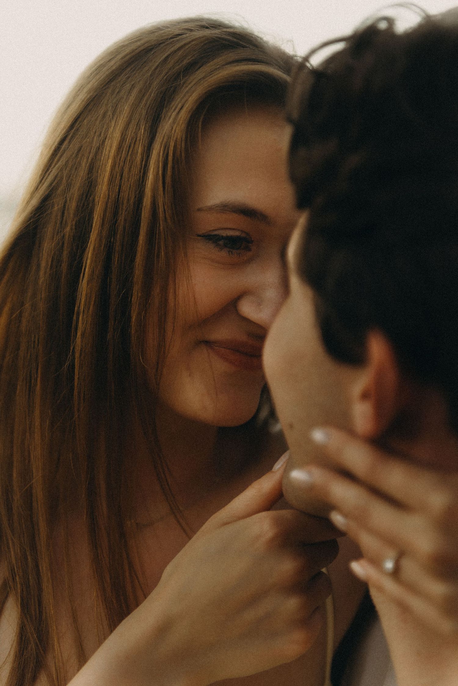 A smiling woman with long brown hair gently holds a man's face close to hers in a romantic moment.