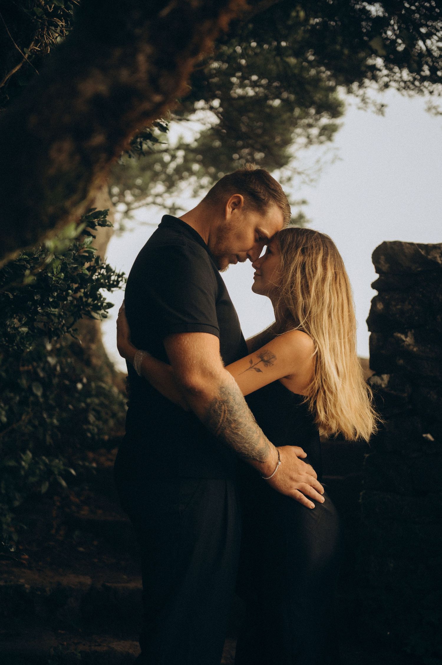 A couple in black outfits share an intimate moment, foreheads touching, surrounded by lush greenery and stone walls.