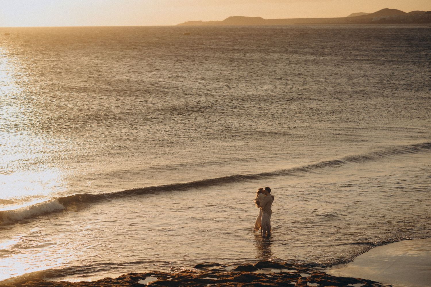 A couple walks along a serene beach at sunset with mountains in the distance creating a romantic silhouette.