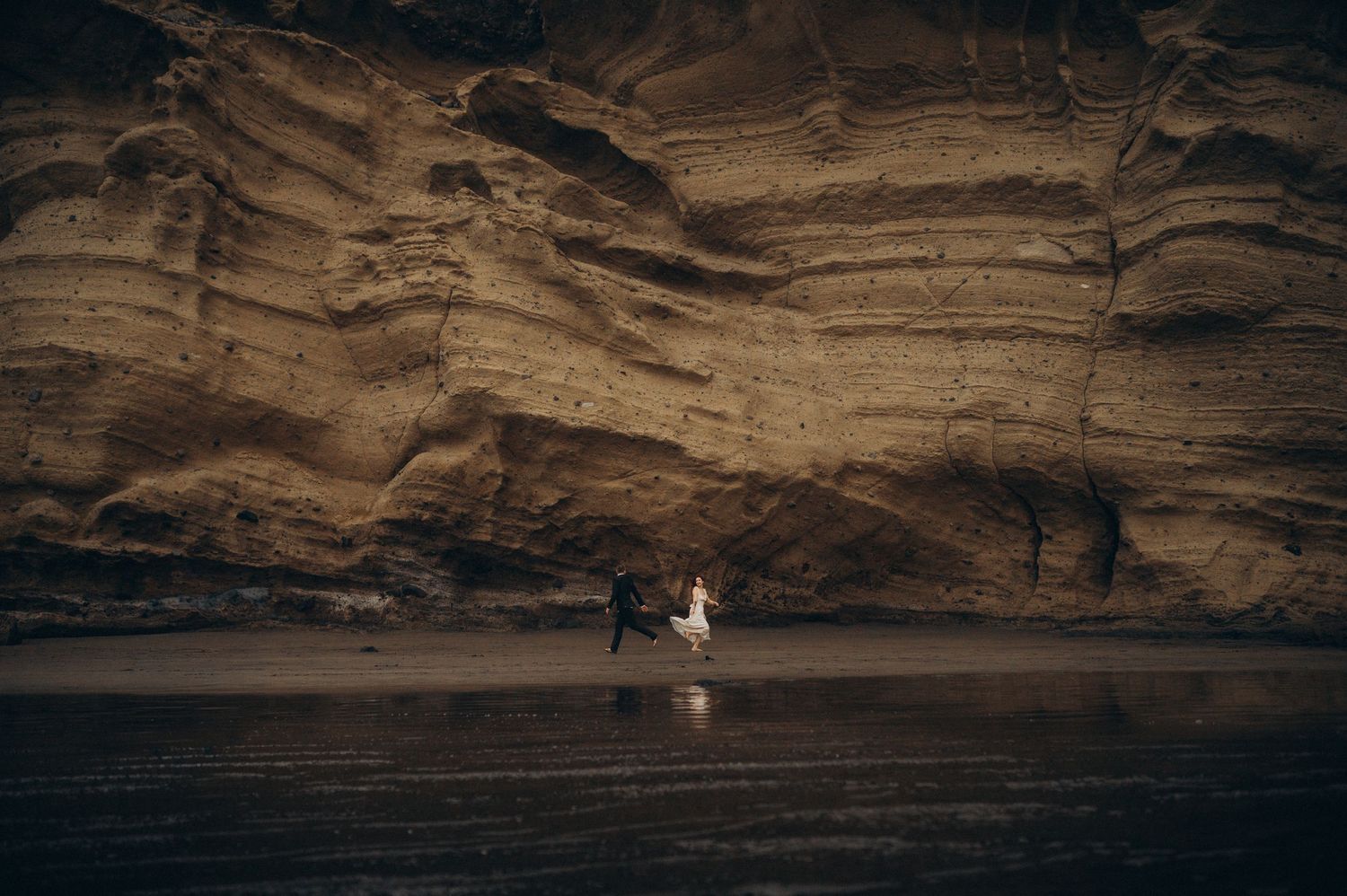 Couple walking on dark beach beneath dramatic layered sandstone cliffs in moody coastal landscape.