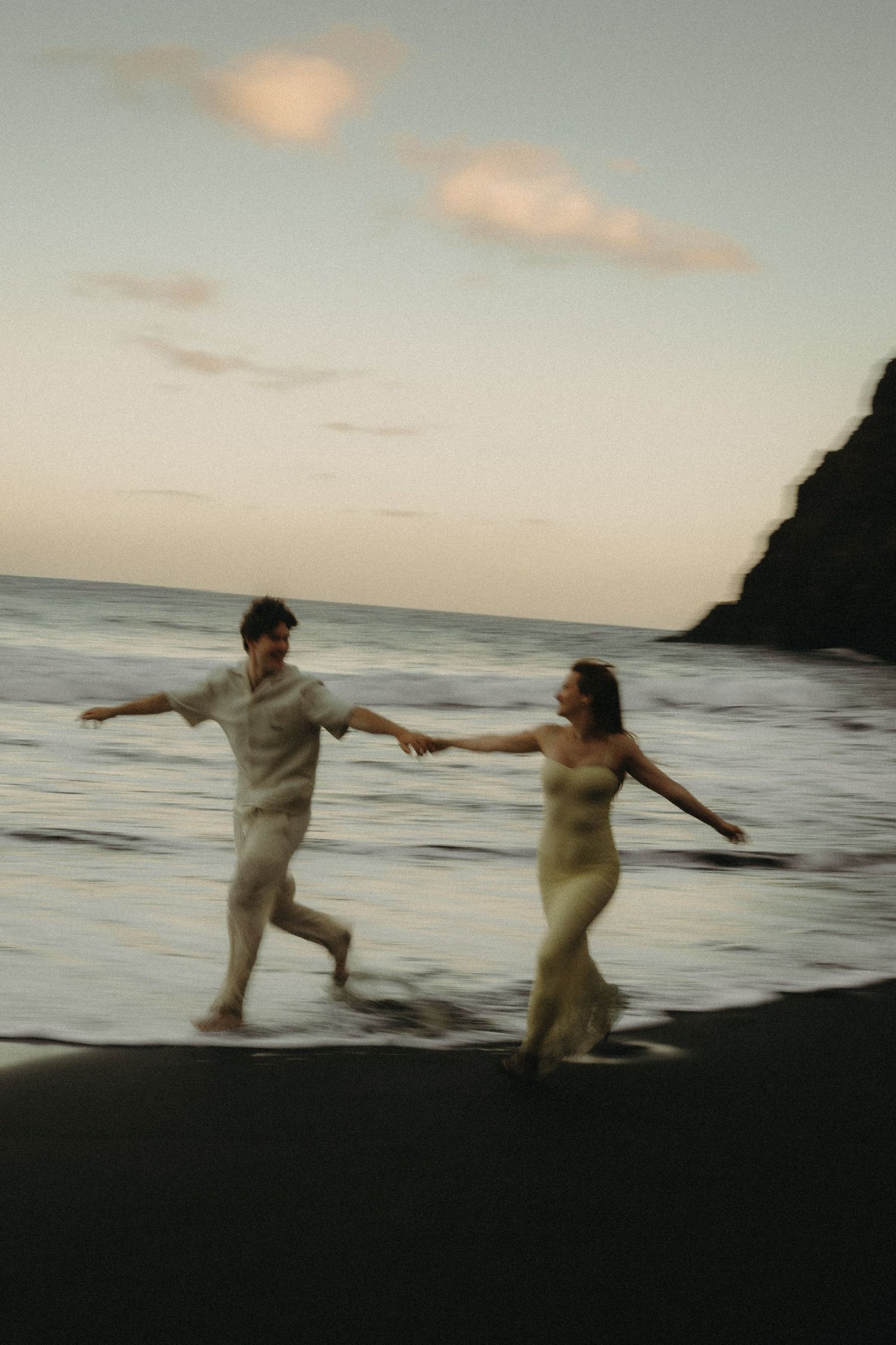 Couple holding hands running along a dark sandy beach at dusk with ocean waves and rocky cliffs in the background.