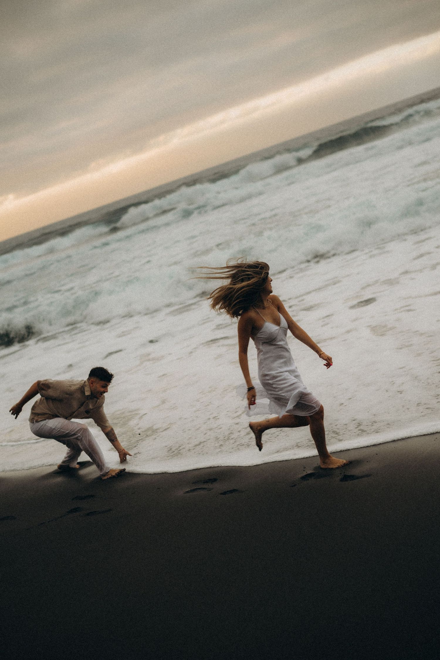 Couple running barefoot on a dark sandy beach at sunset, woman in white dress with hair blowing, waves crashing behind them.