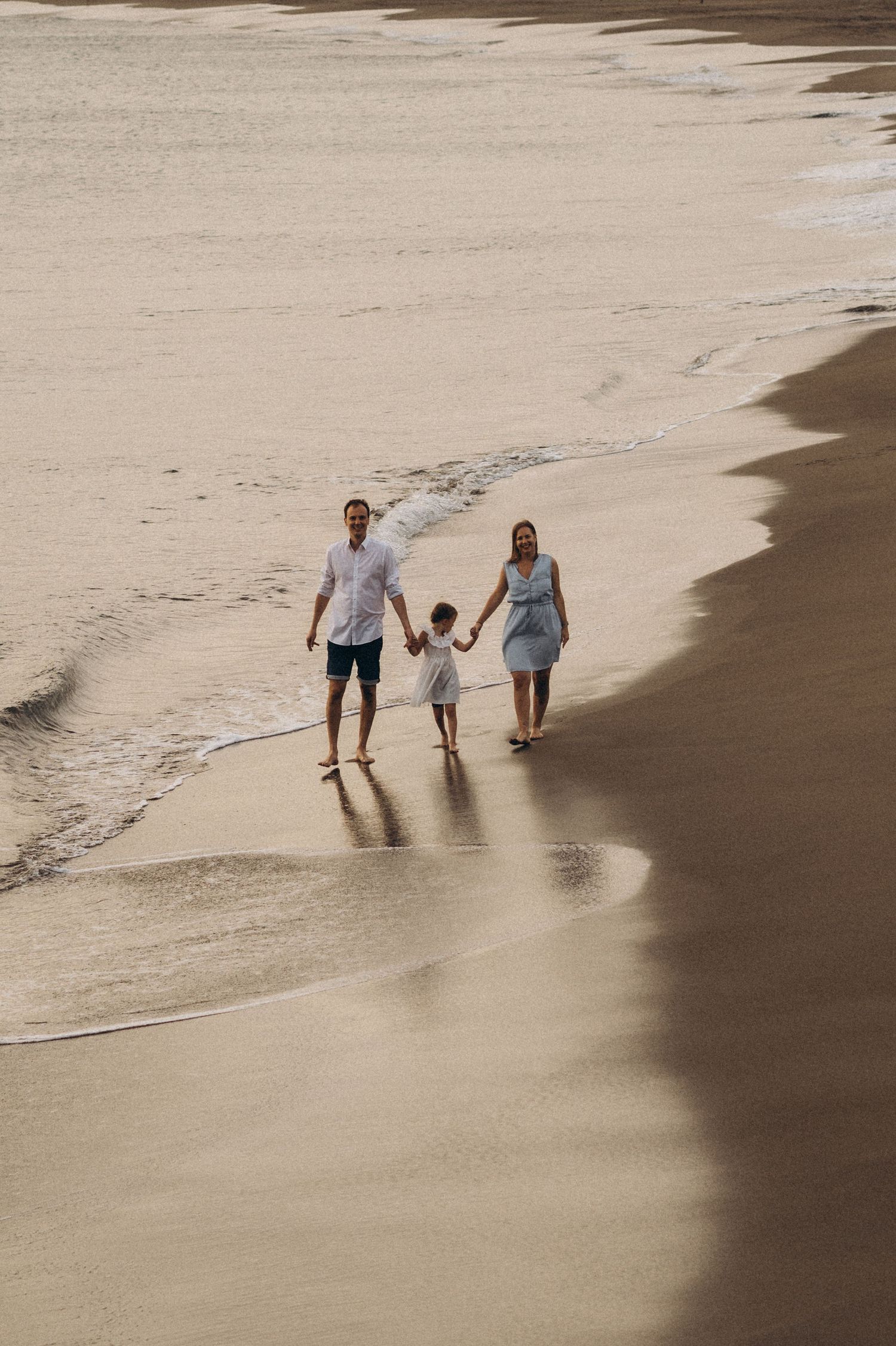 A family walks along a sandy beach shoreline holding hands as waves gently lap at their feet.