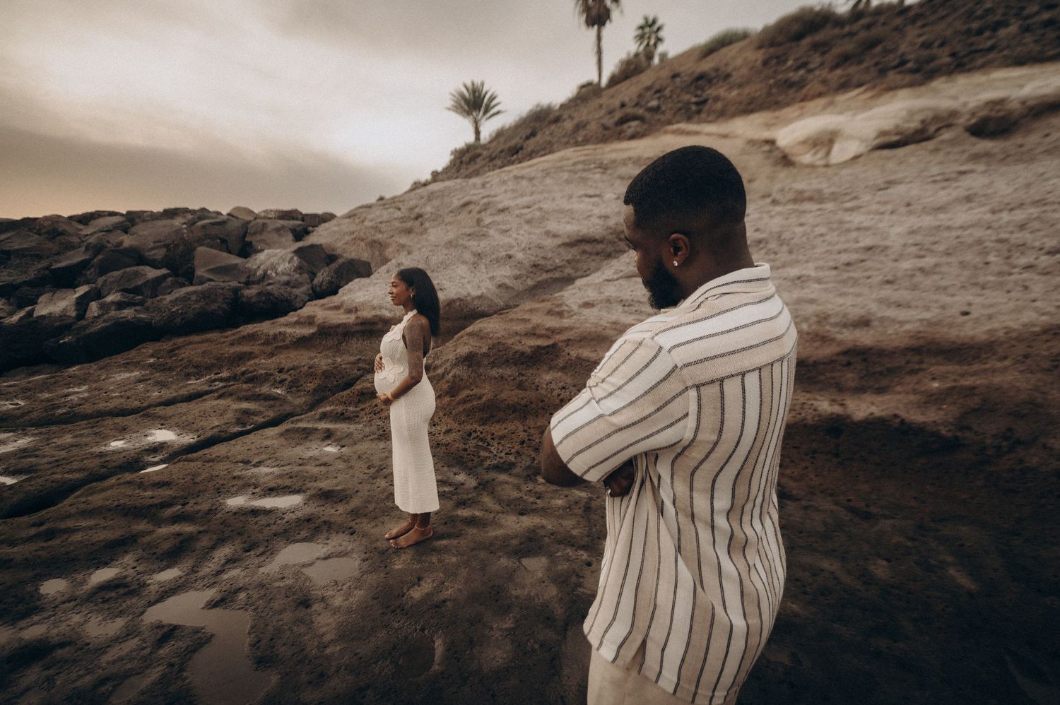 Man admiring pregnant wife in foreground with cliffs behind, Playa Fañabé