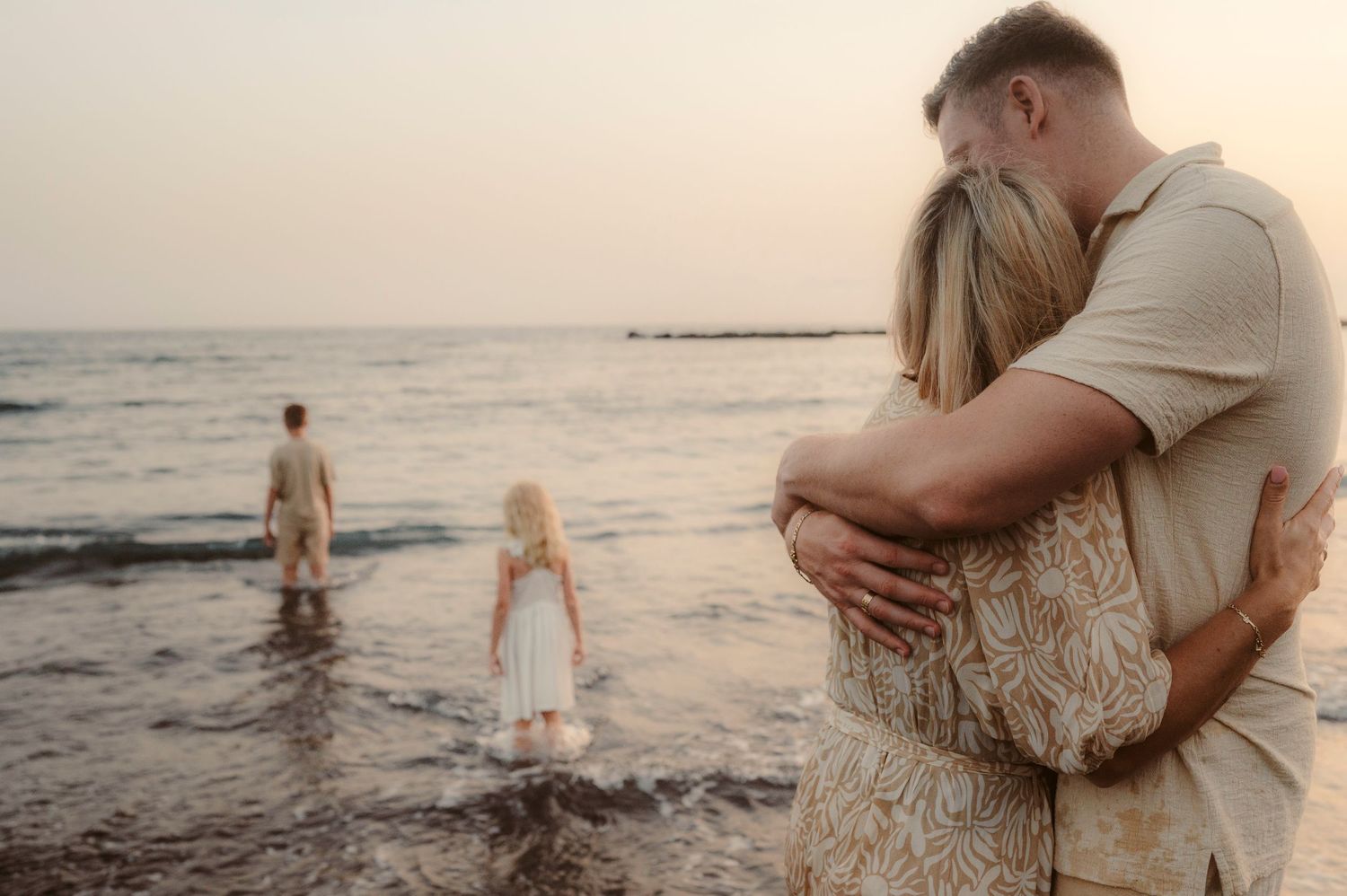Parents hugging while watching their children play in ocean at sunset, Playa del Duque