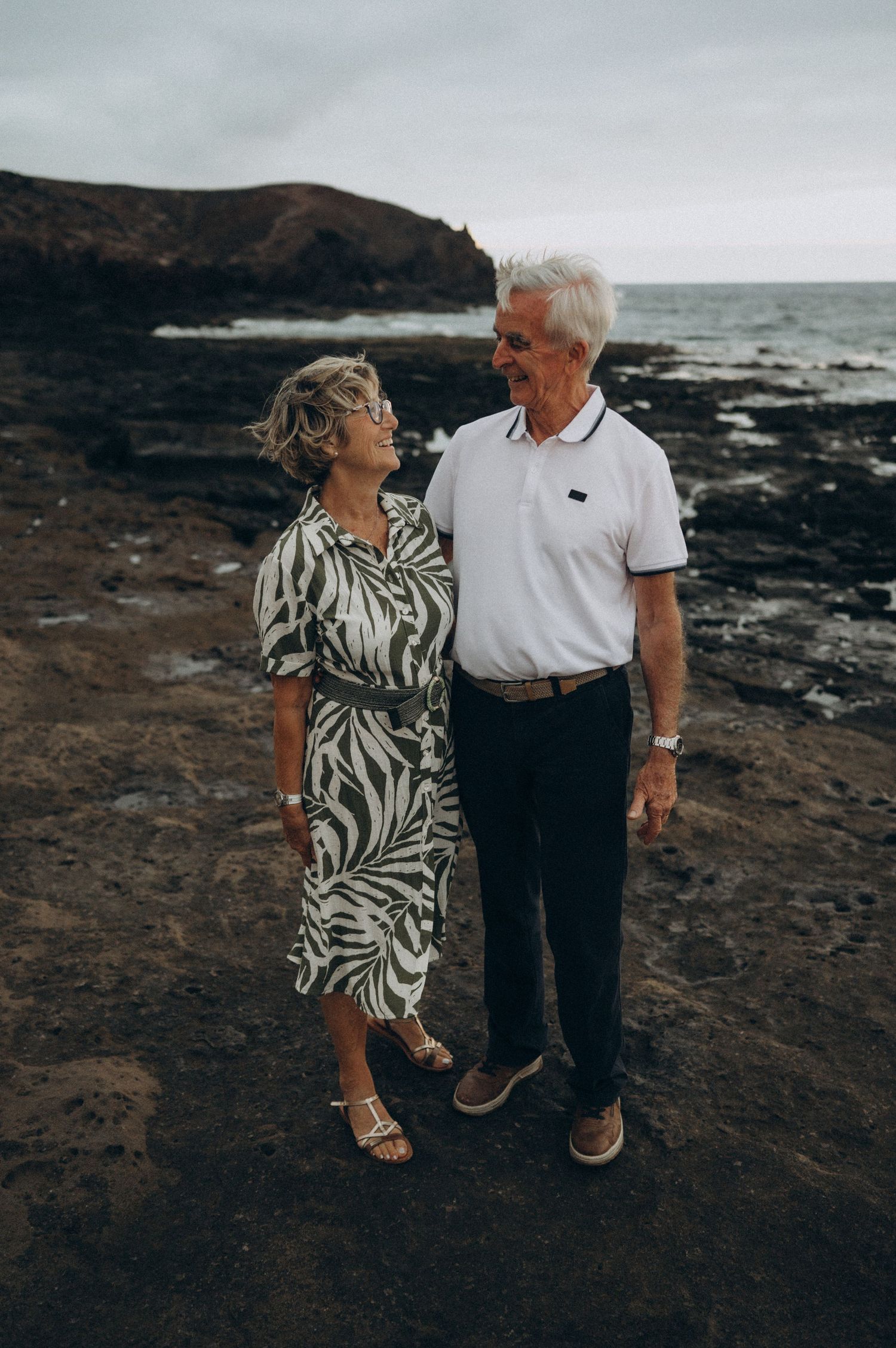 Senior couple holding each other on rocky ground with ocean and cliffs at Los Refugios